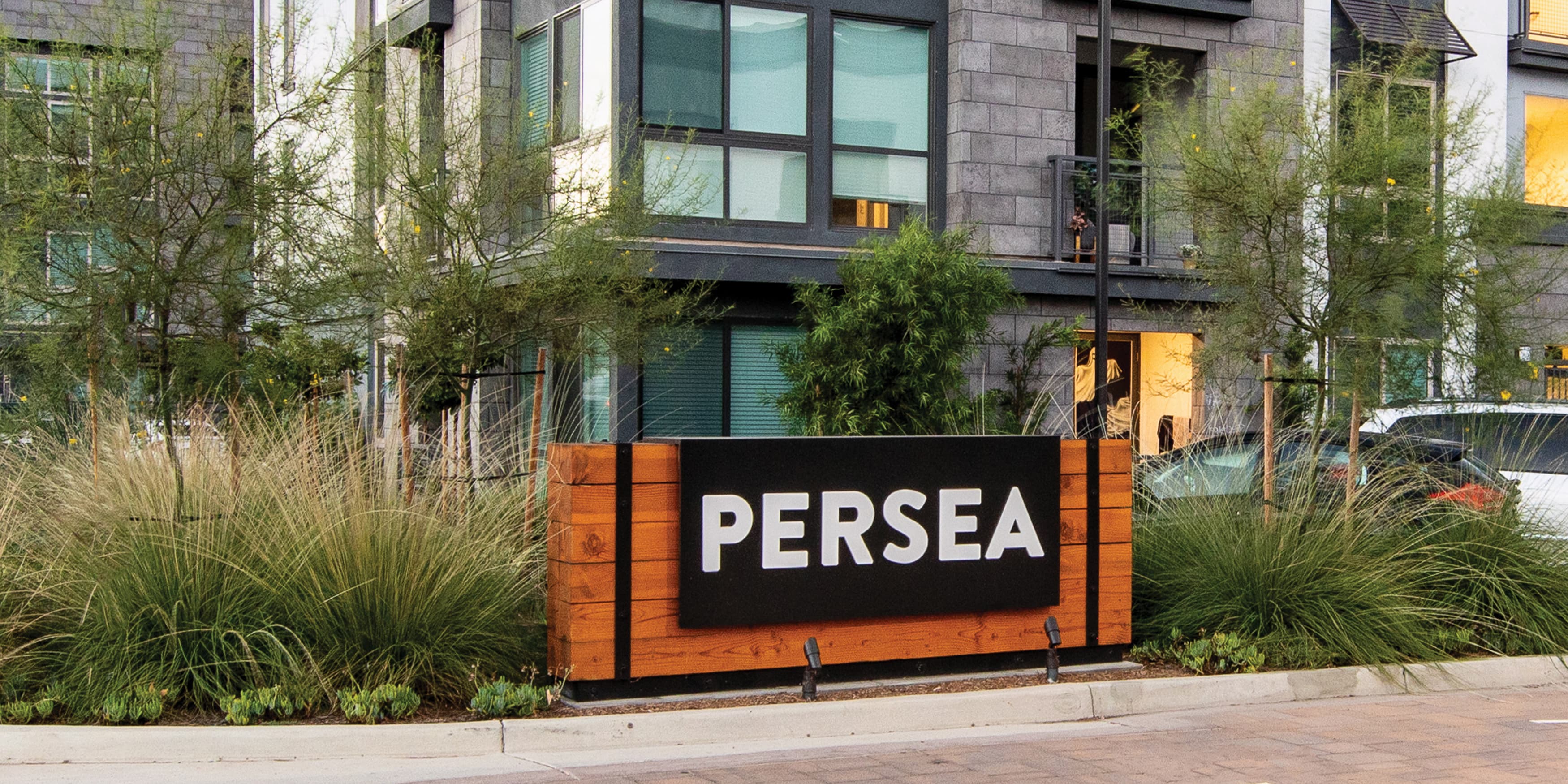 Horizontal rustic modern Persea identity monument sign with horizontal stacked wood posts under a black metal cap with the Persea identity in white on top and exposed black fasteners. Signage and wayfinding for Persea in San Diego, California by RSM Design