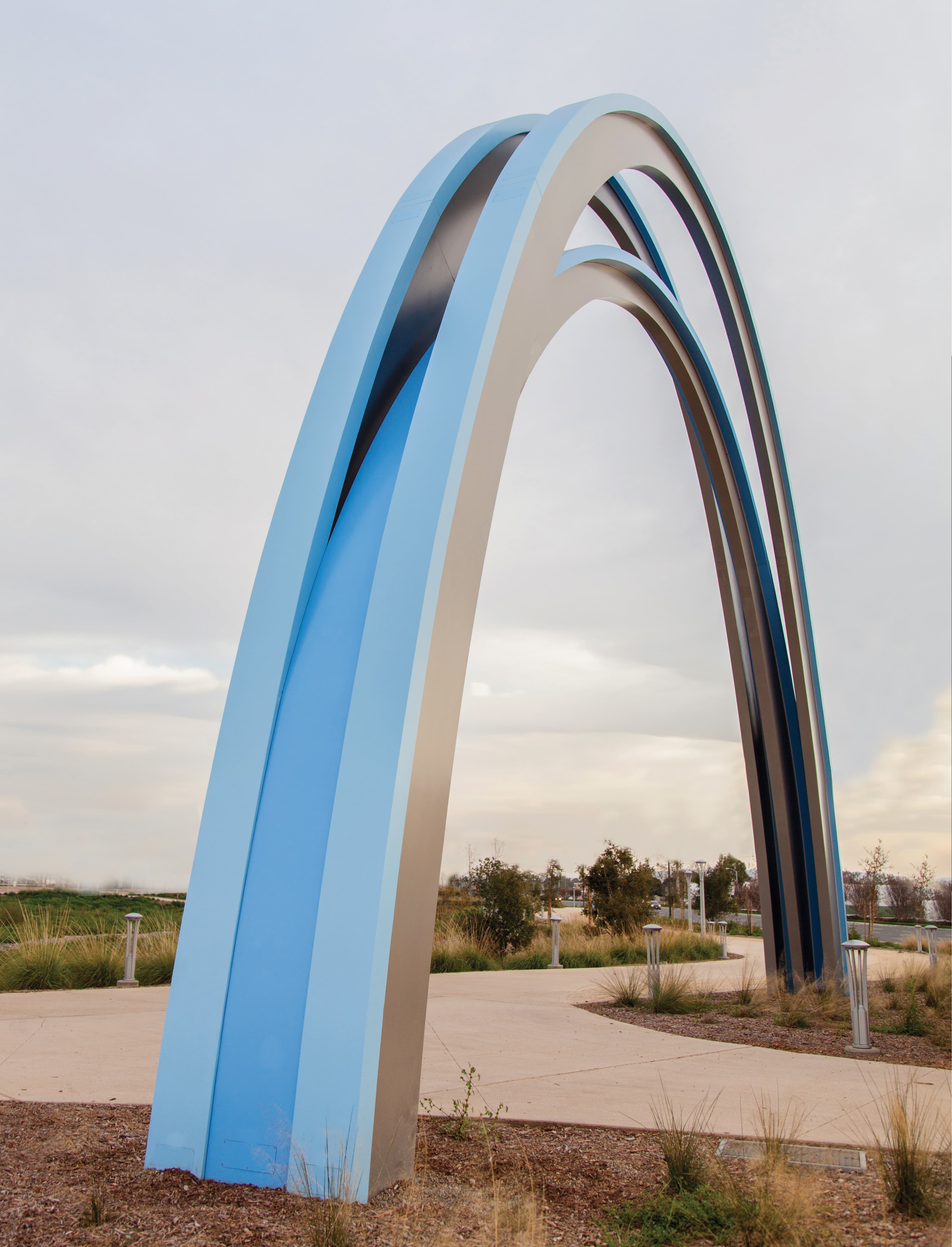 Full view of a tall sculptural archway with layered blue and cream bands, standing over a paved walking path lined with native grasses and lampposts, with trees and an overcast sky in the background.
