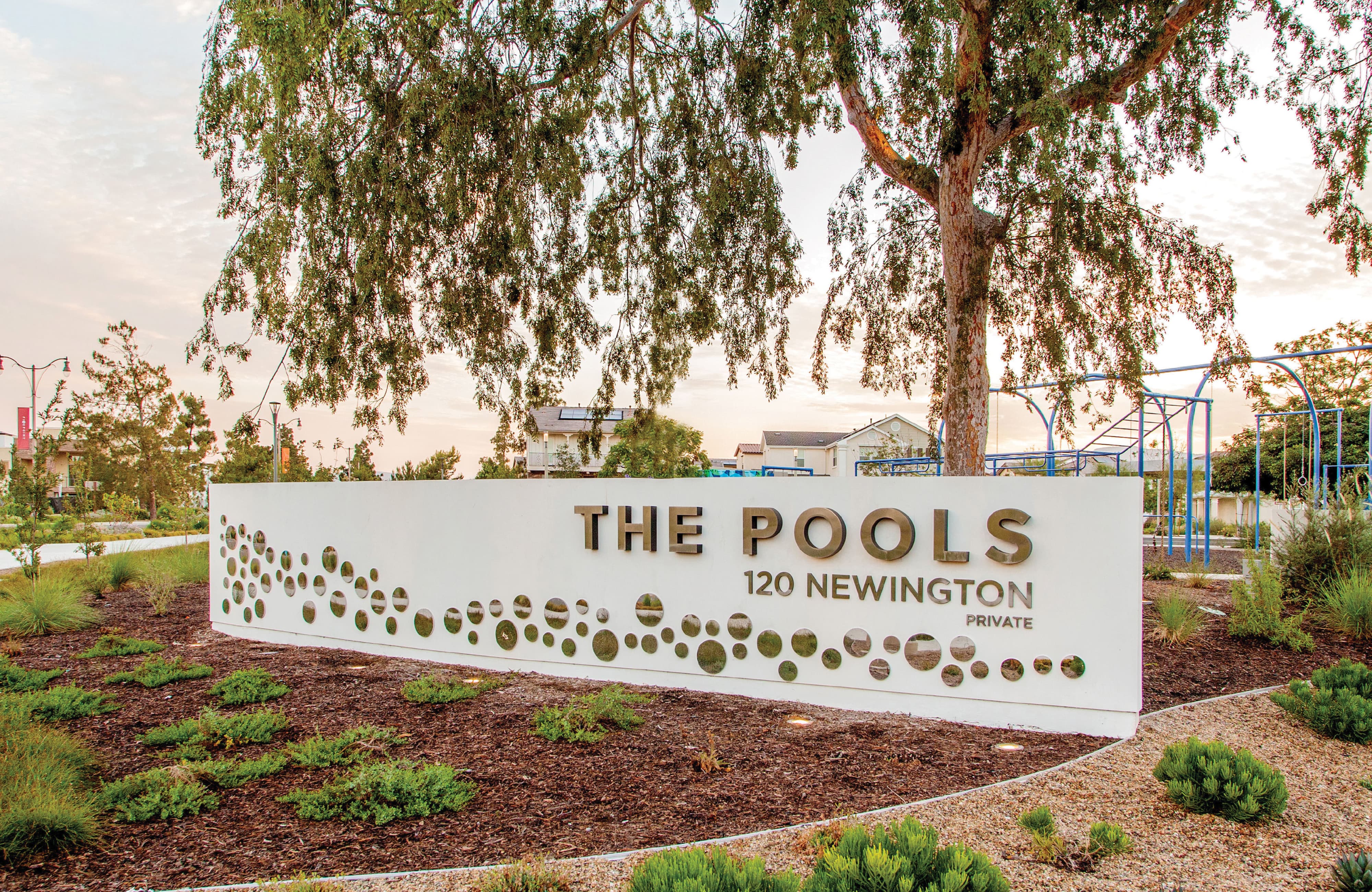 Monument sign reading 'The Pools, 120 Newington, Private' in gold lettering on a white wall with a decorative circular cutout pattern, set beneath a tree with a playground and residential homes in the background.