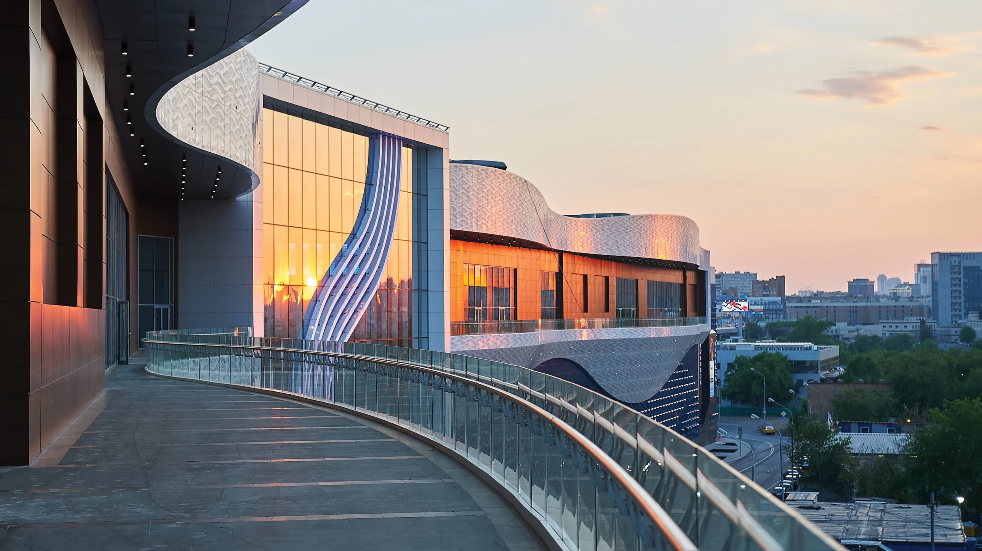 Elevated exterior walkway at sunset along a large-scale civic or cultural building facade featuring sweeping architectural forms, large-format decorative graphic panels, and a curvilinear glazed railing with city views — RSM Design experiential graphics, specialty graphics, placemaking, and environmental graphic design for civic, cultural, and mixed-use destinations.