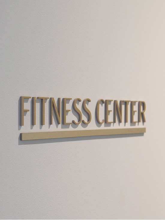 The Monarch at Hall Park amenity room identification signage — dimensional brushed bronze "Fitness Center" sign with rule bar detail on white wall, interior wayfinding design by RSM Design