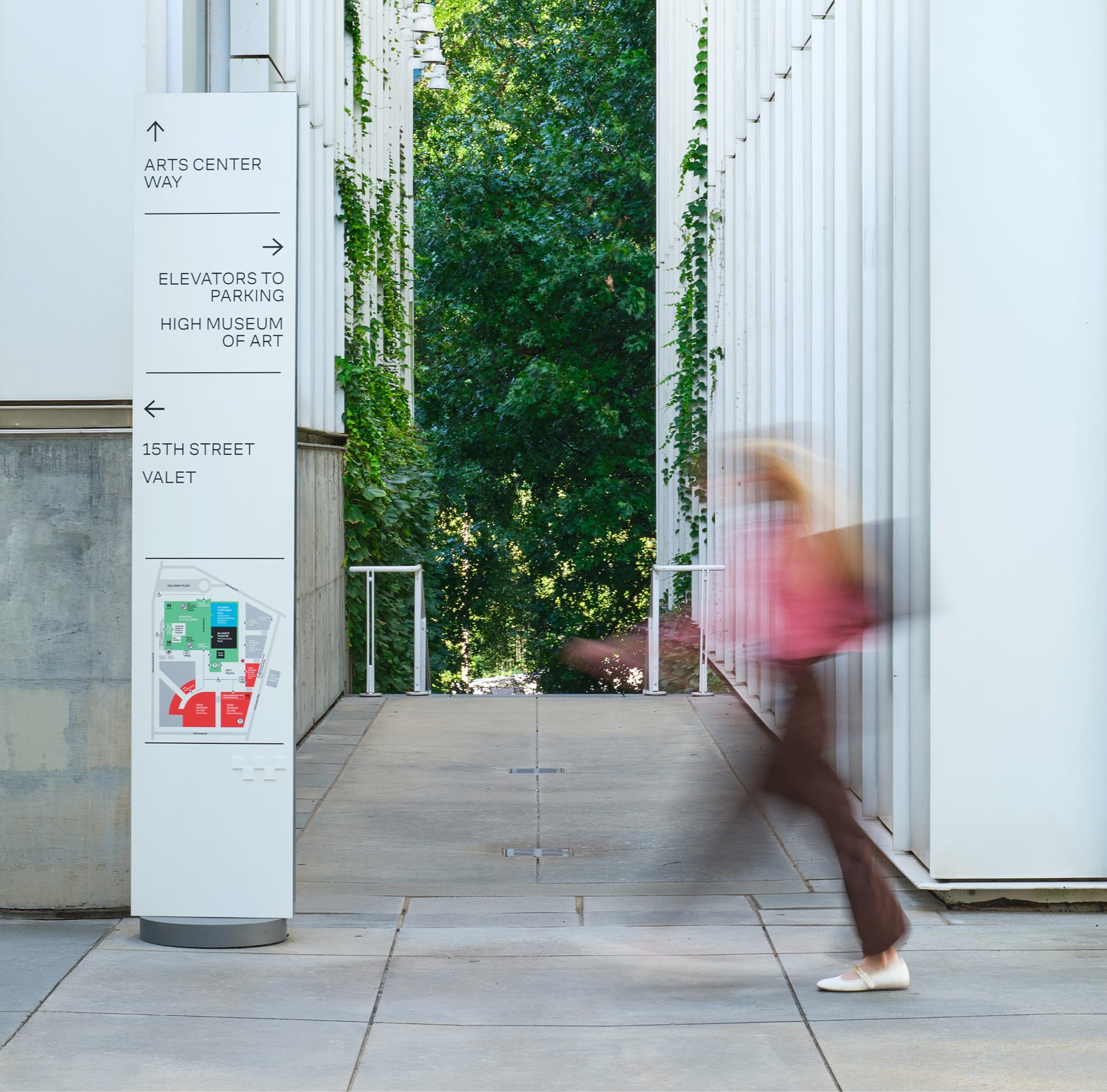 Directional pylon sign with campus map guiding visitors to Arts Center Way, elevators to parking, the High Museum of Art, and 15th Street valet, set beside a colonnaded walkway at the Woodruff Arts Center – campus wayfinding and environmental graphic design by RSM Design.