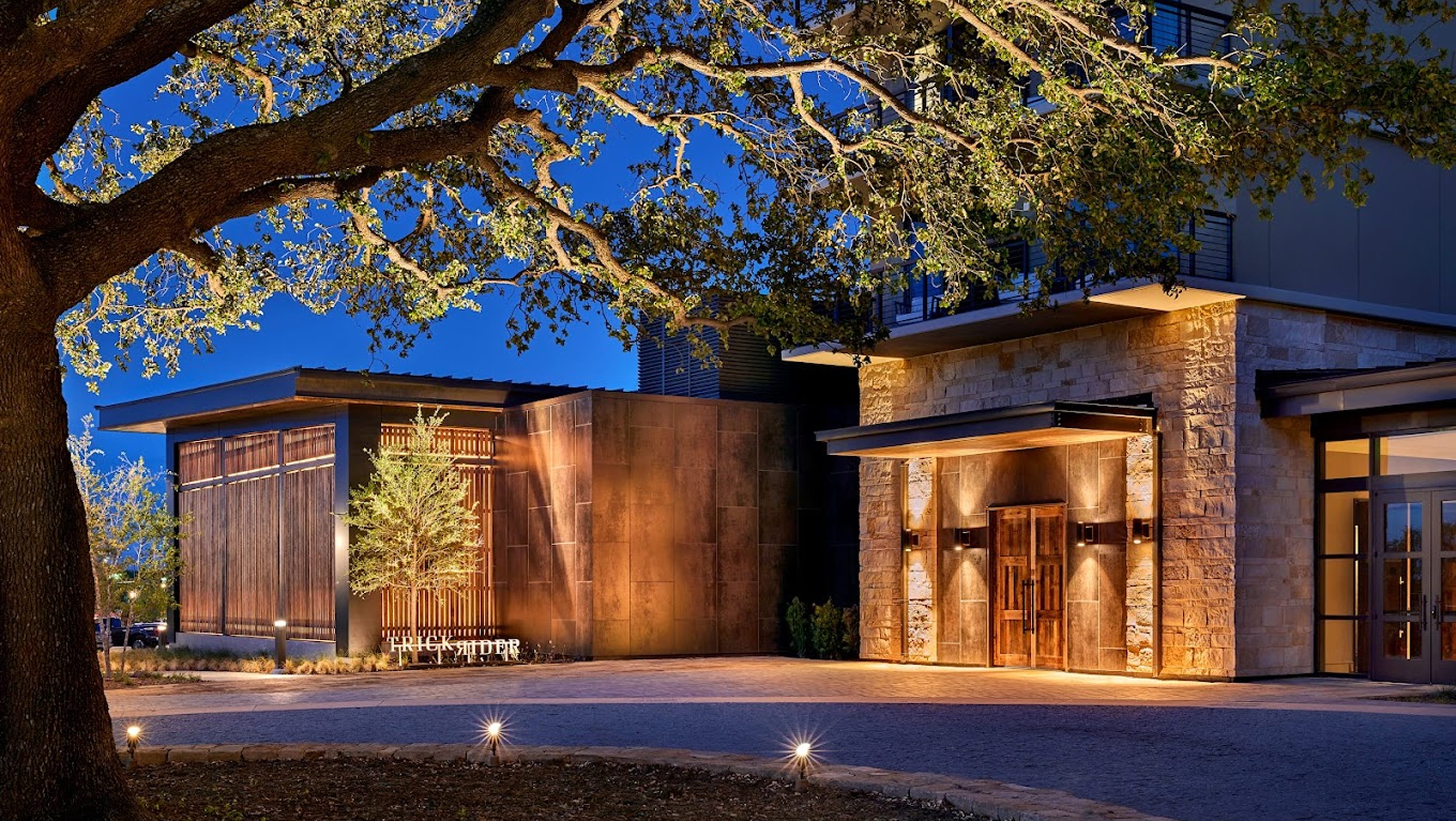 Trick Rider restaurant entrance with illuminated stone facade, wood doors, and patinated metal paneling at dusk beneath oak tree branches, Omni PGA Frisco Resort, environmental graphic design by RSM Design.