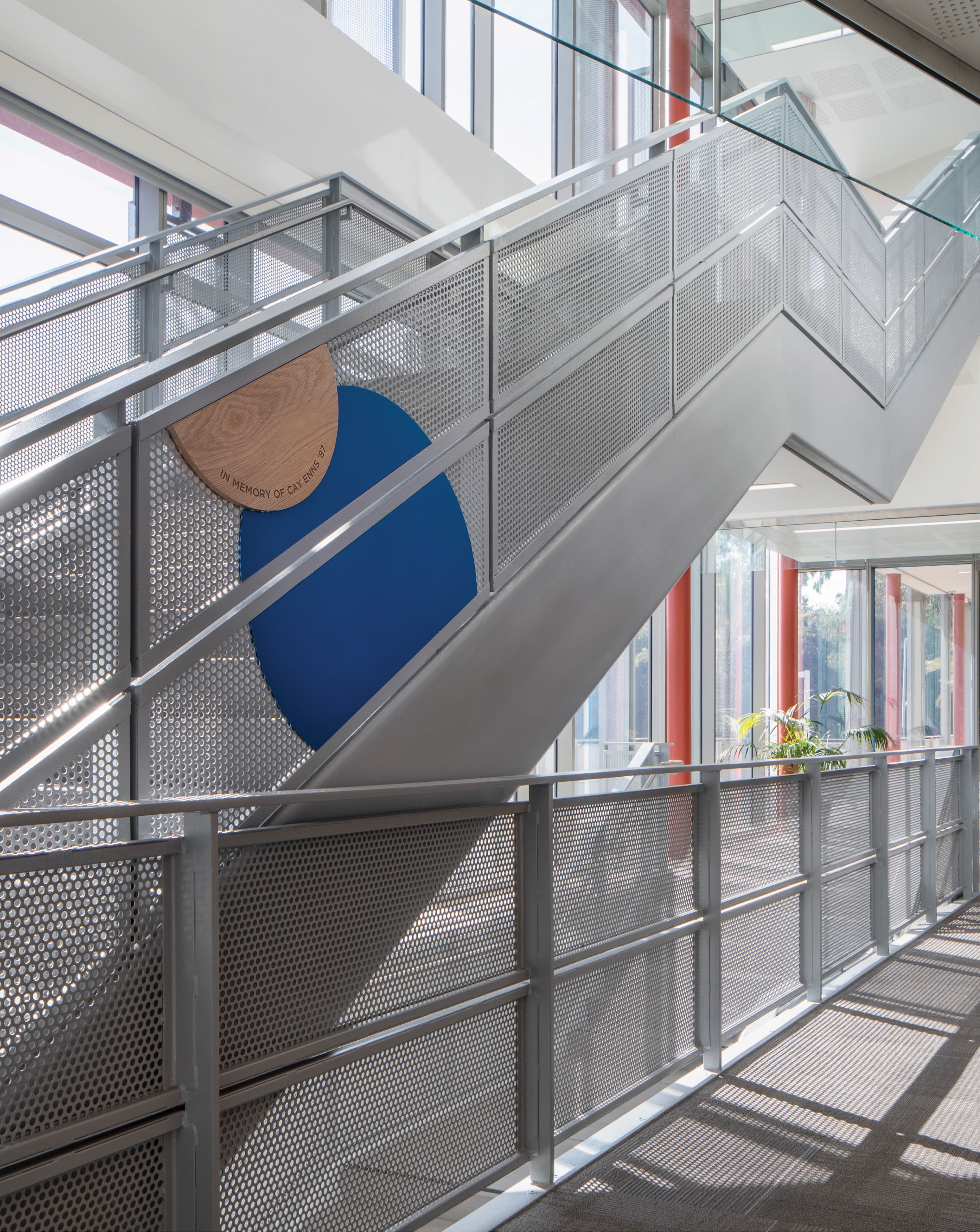 An interior stairwell of an educational campus building featuring a large-scale donor recognition medallion composed of a natural wood half-circle and cobalt blue geometric form mounted on a perforated metal stair panel, inscribed "In Memory of Cay Enns '87," with dappled natural light casting geometric shadows across the industrial railings and glazed corridor beyond — showcasing RSM Design's donor recognition and specialty graphics and art programs services integrated thoughtfully into the architecture of the education market