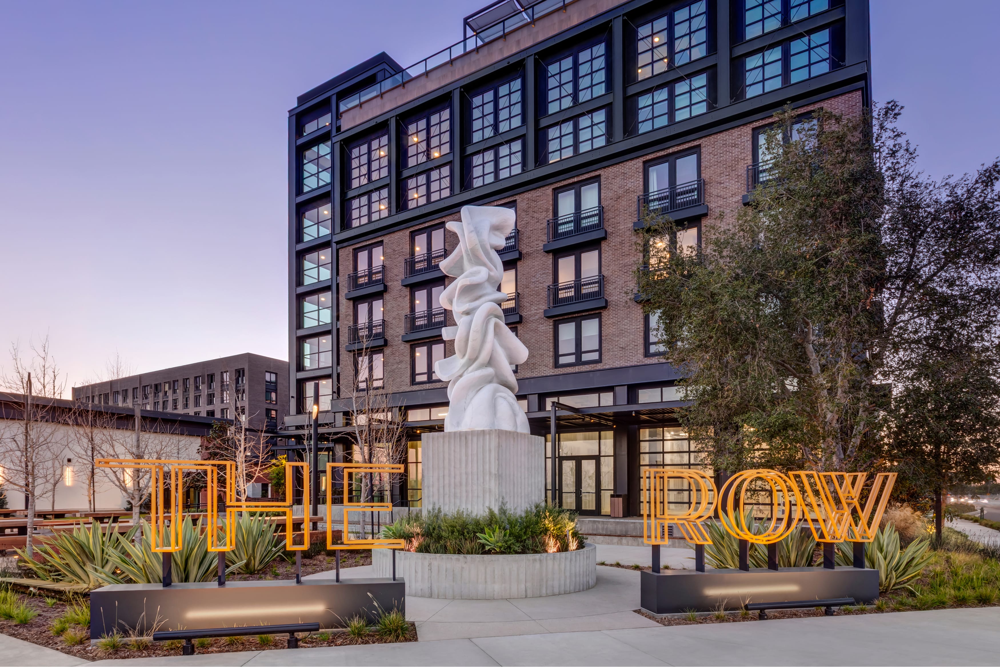 Illuminated amber "THE ROW" channel letter monument sign flanking a white sculptural art piece at dusk, with brick and black-framed apartment building behind at The Row at Red Hill – placemaking and environmental graphic design by RSM Design.