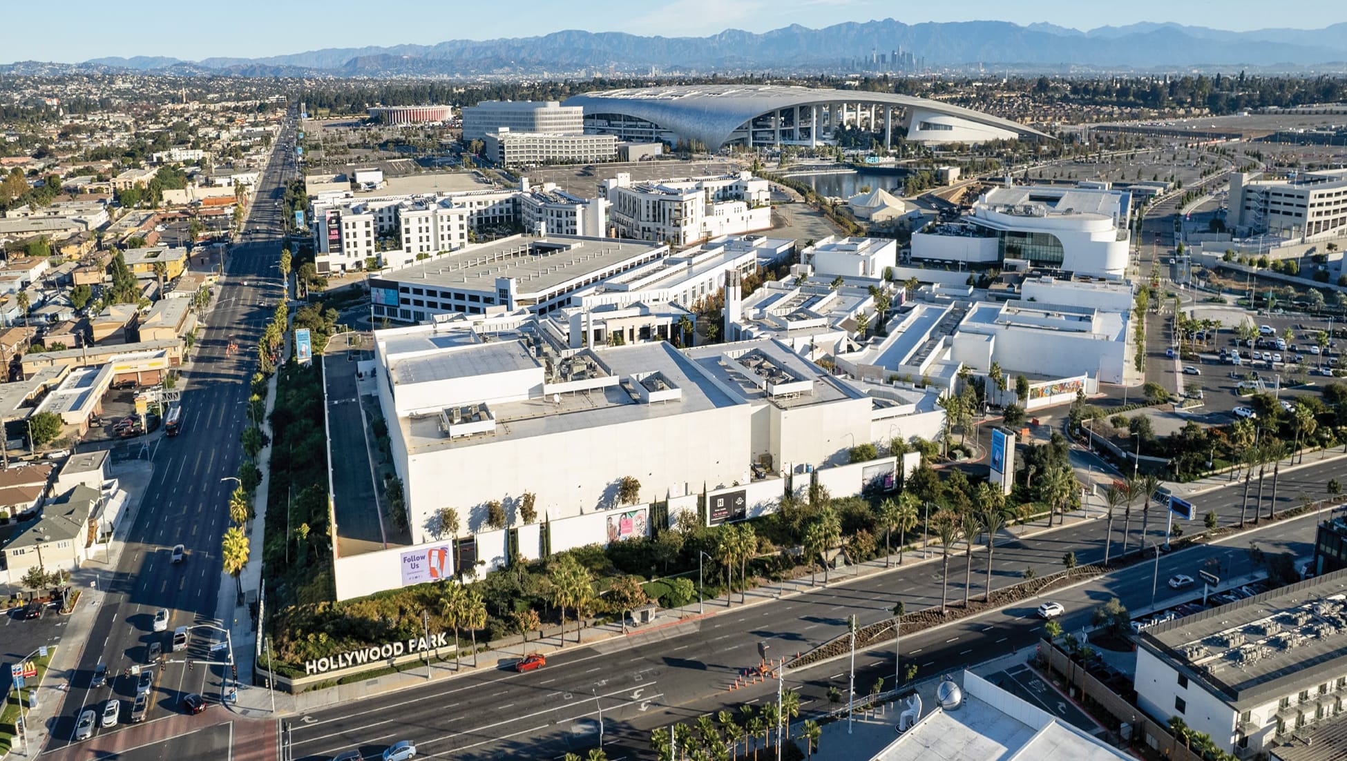 Wide aerial view of Hollywood Park mixed-use retail and entertainment complex in Inglewood, California with monument sign, SoFi Stadium, Kia Forum, residential buildings, palm-lined boulevards, and Los Angeles skyline and mountains in background