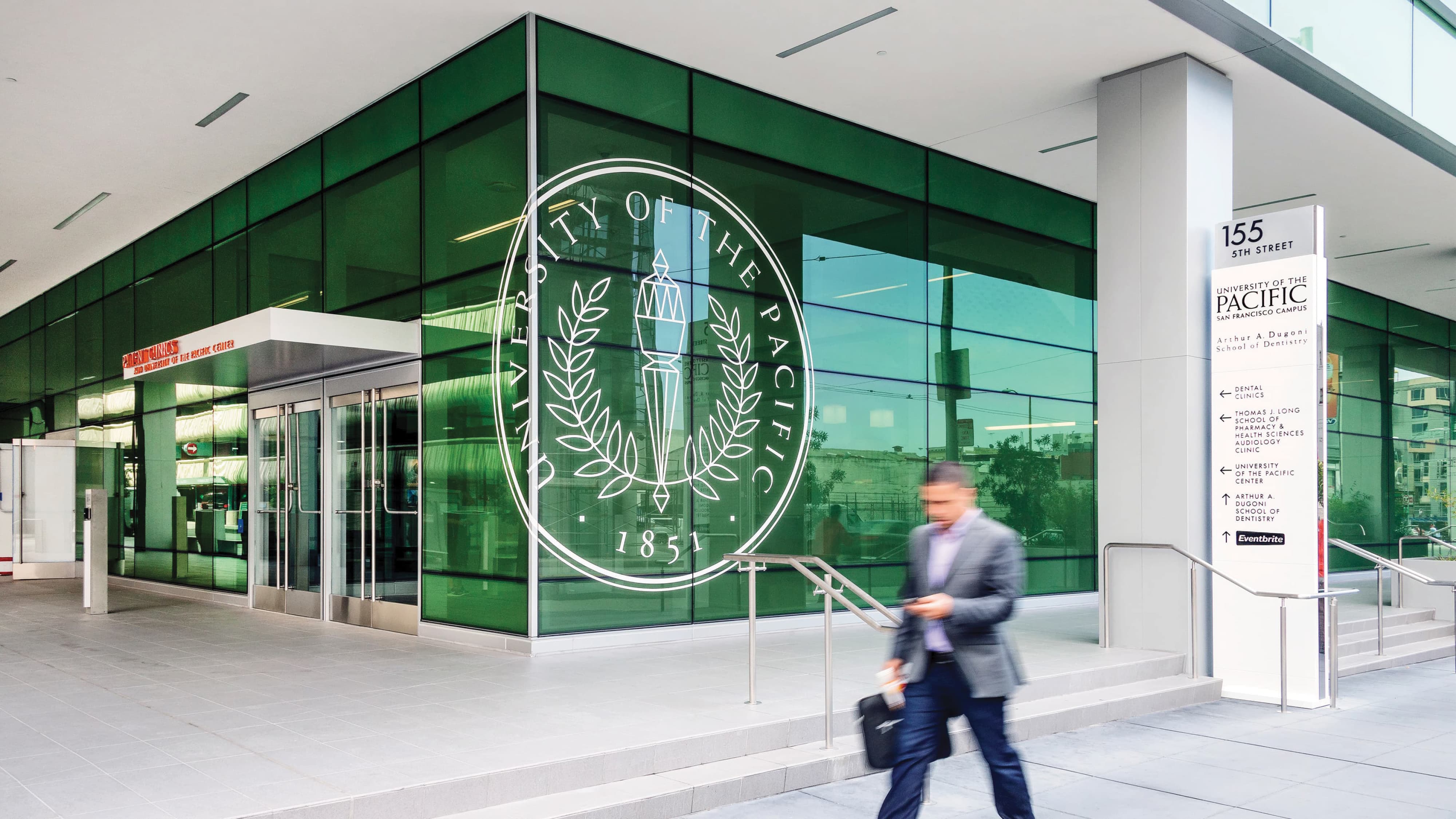 University of the Pacific San Francisco Campus exterior building entry featuring a large-scale frosted glass university seal graphic established in 1851 on deep green glazing, paired with a multi-destination pedestrian wayfinding pylon directing visitors to dental clinics, pharmacy, health sciences audiology clinic, and Arthur A. Dugoni School of Dentistry — RSM Design branding and visual identity, wayfinding and signage design, master sign programs, specialty graphics, and environmental graphic design for education, health and wellness, and civic destinations.