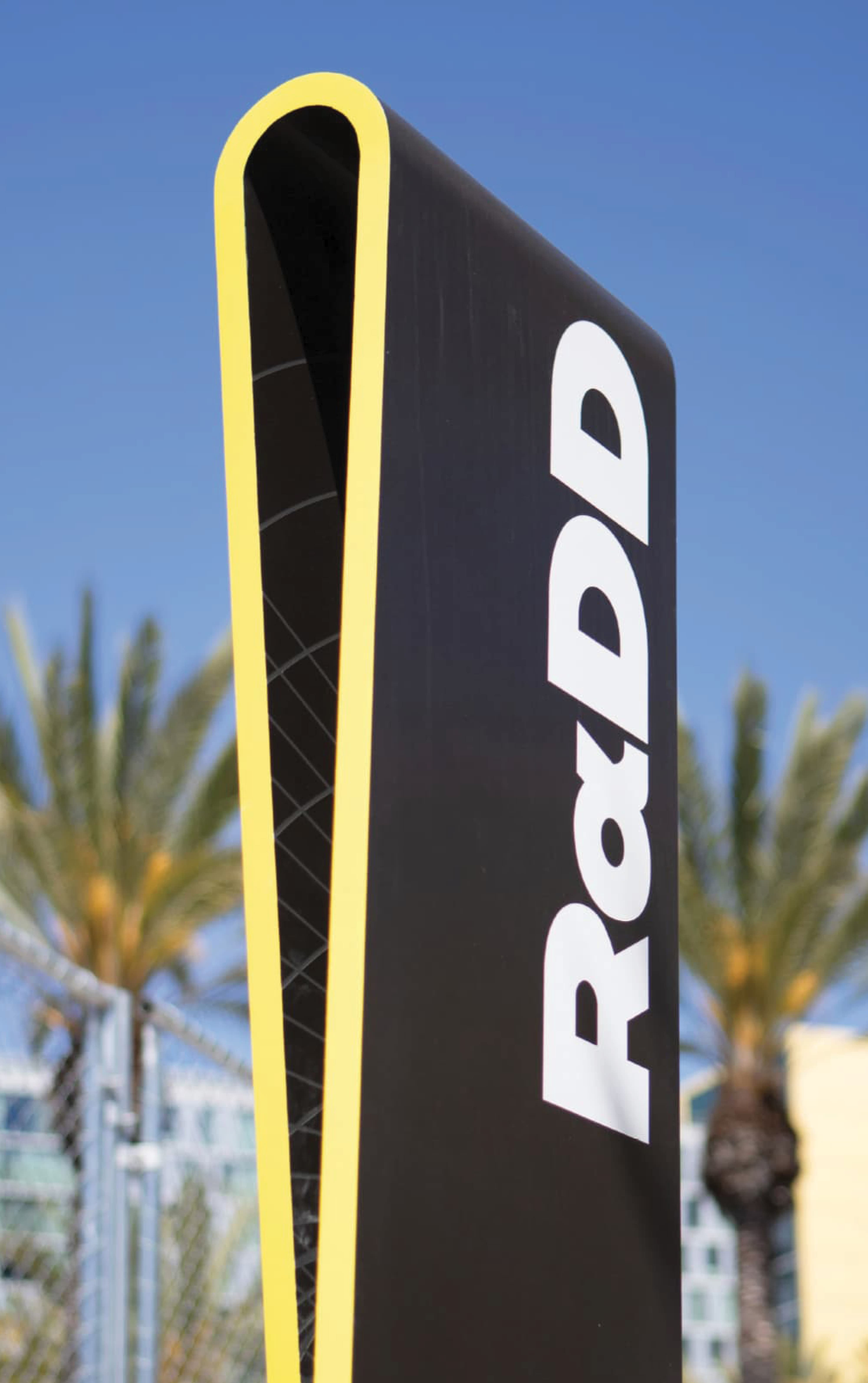 Tall black monument sign with a curved yellow-edged channel and 'RαDD' lettering in white running vertically down the side, set against a blue sky with palm trees and buildings in the background.