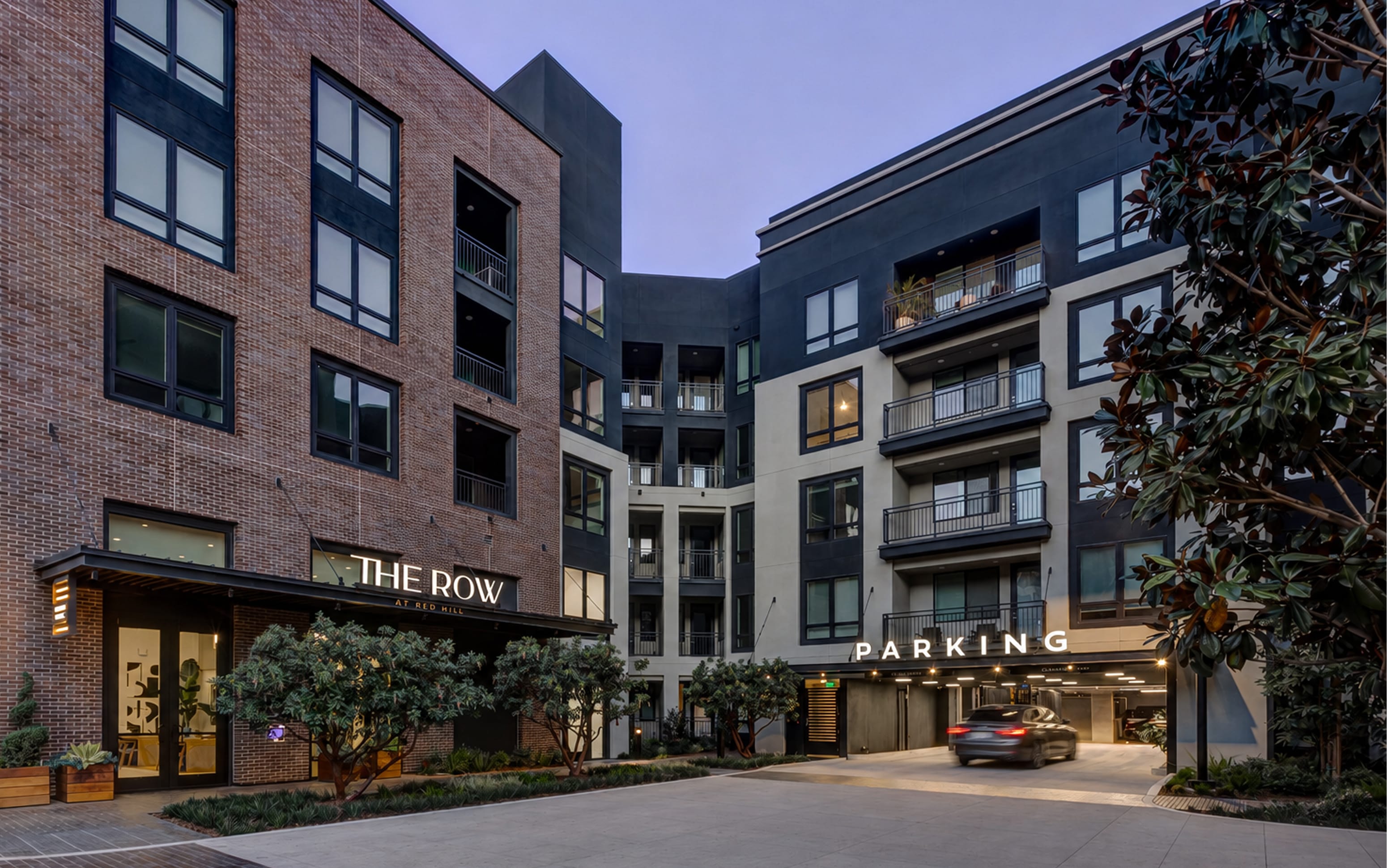 Illuminated "THE ROW AT RED HILL" building identification sign and backlit "PARKING" wayfinding sign on brick and dark-toned apartment buildings at dusk – placemaking and environmental graphic design by RSM Design.