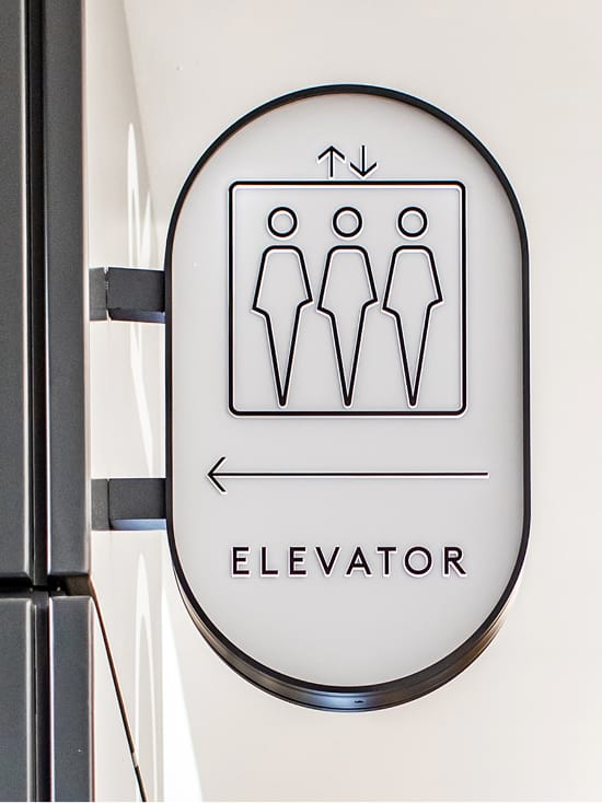 Ovation Hollywood elevator directional blade sign with custom illustrated pictogram, black oval backlit frame, and directional arrow on white wall