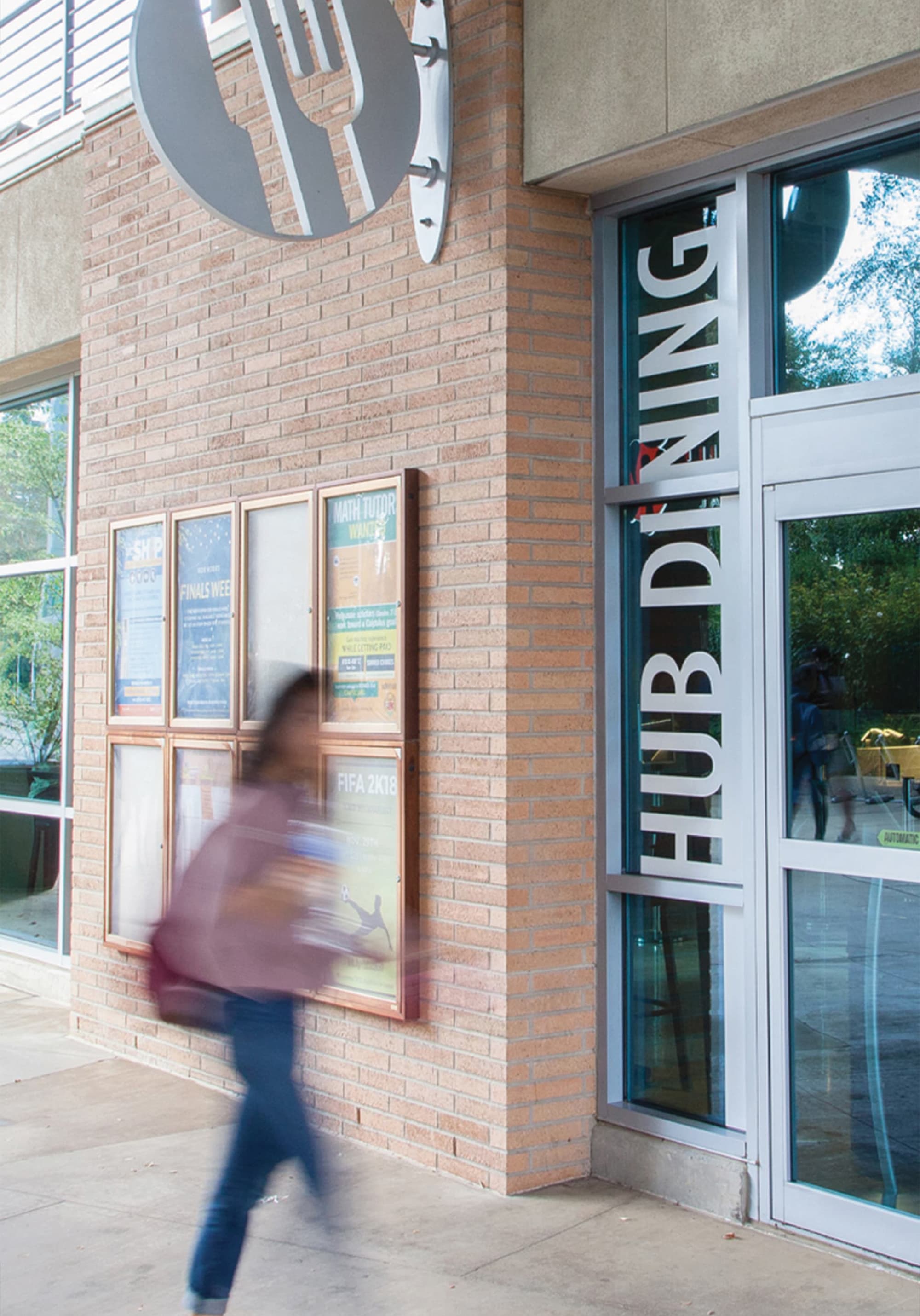 Brick building corner with a circular fork-and-plate icon above a bulletin board of flyers, and vertical 'HUB Dining' text on a glass entrance to the right, as a student walks past in motion blur.