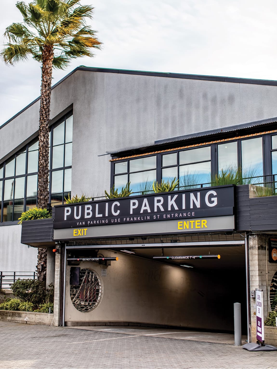 Jack London Square public parking garage entrance and exit with bold yellow signage, decorative nautical medallion, rooftop planters, and palm tree in Oakland, California