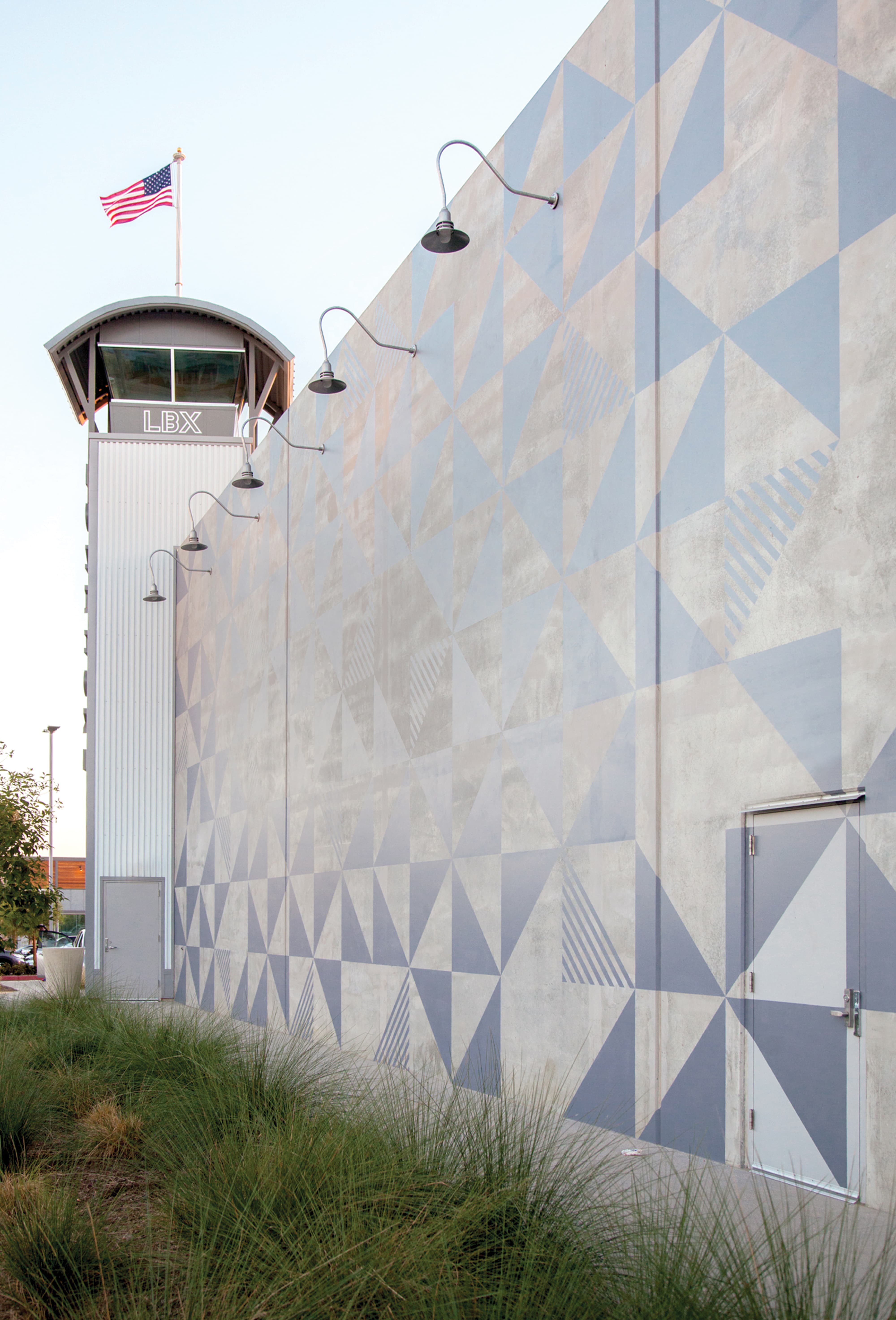Angled view of a building's exterior wall painted with a gray geometric triangle pattern, leading toward a control-tower-style structure labeled 'LBX' topped with an American flag, with wall-mounted lights and native grasses in the foreground.