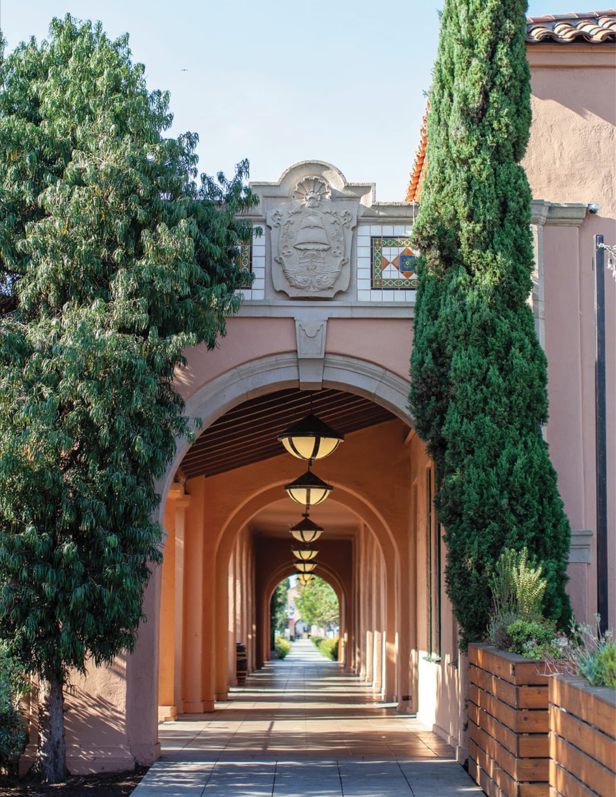 Spanish Colonial style covered walkway with terracotta-colored arches, hanging lantern lights, and decorative tile crest above the entrance, framed by tall cypress and shade trees.