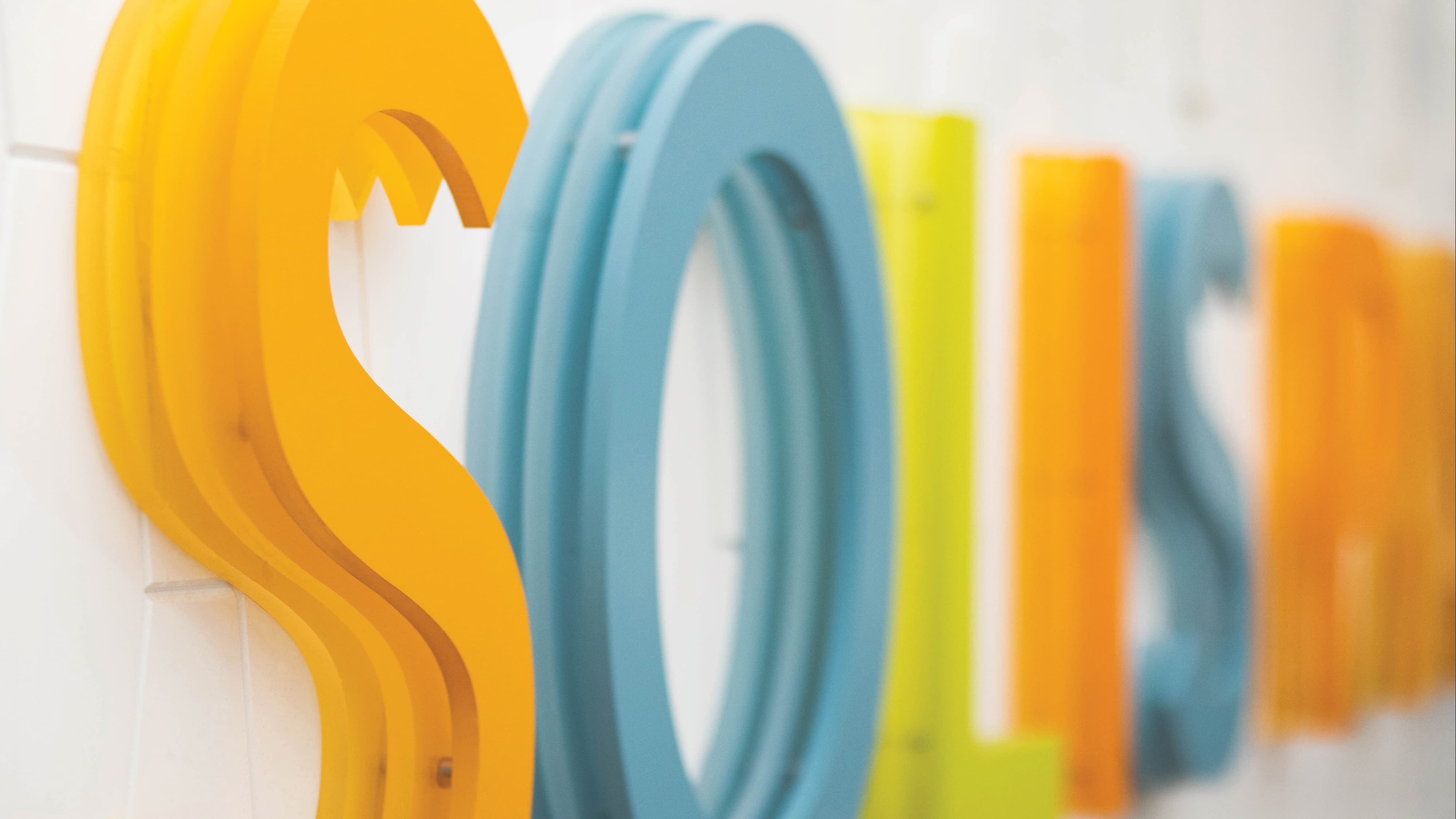 Close-up detail of large-scale dimensional letters spelling "SOLIS" in layered yellow, teal, lime green, and orange painted forms mounted on a white wall — highlighting RSM Design's specialty graphics, branding and visual identity, and experiential graphics and branded environments work applied to a health and wellness or residential community environment