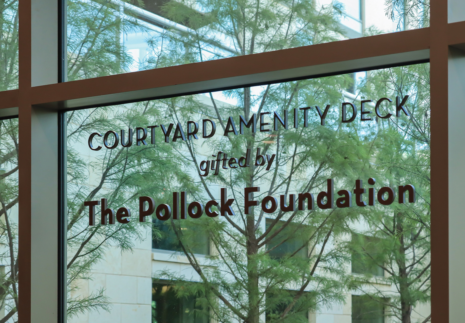 Glass window signage reading 'Courtyard Amenity Deck, gifted by The Pollock Foundation,' with feathery trees and a building courtyard visible through the glass.