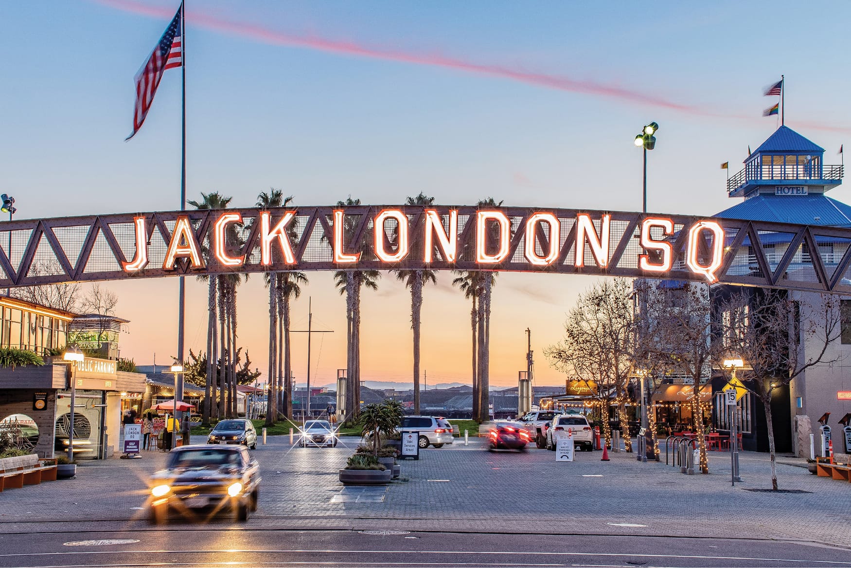 Illuminated Jack London Square entrance sign at sunset in Oakland, California, featuring neon “Jack London SQ” lettering, palm trees, waterfront views, and active street scene near the marina and restaurants.