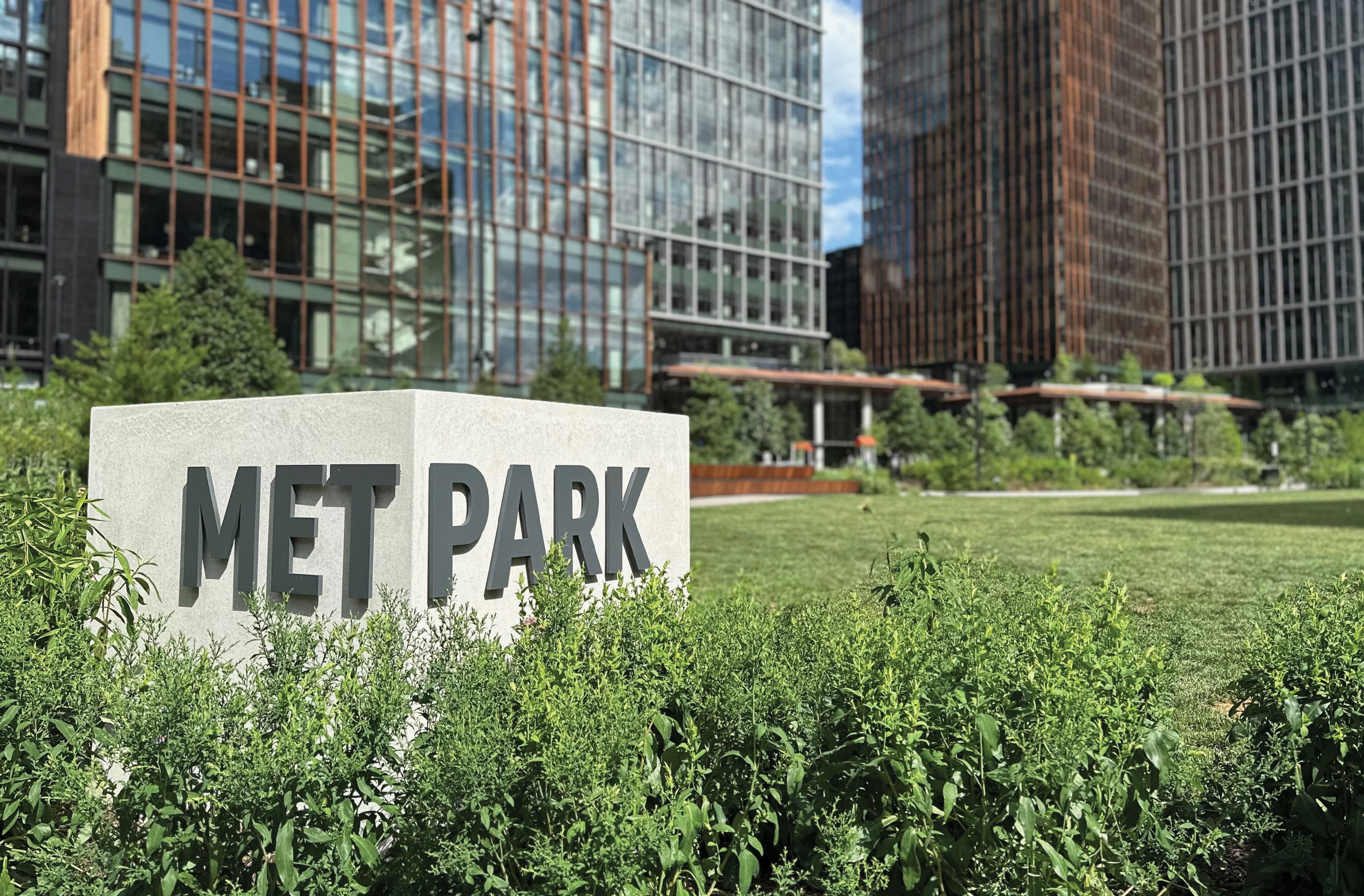 
This image depicts the "MET PARK" monument sign situated within a lush green space. The sign is made of concrete with bold, black lettering, and it stands amidst dense vegetation. In the background, modern high-rise buildings with extensive glass facades are visible, blending with the greenery of the park. The buildings feature a mix of vertical and horizontal design elements, enhancing the contemporary urban aesthetic. The well-maintained lawn and surrounding plants create a serene atmosphere, making the park an inviting spot within the urban landscape. The image captures the integration of natural elements within a bustling city environment, showcasing a balance between modern architecture and green spaces.