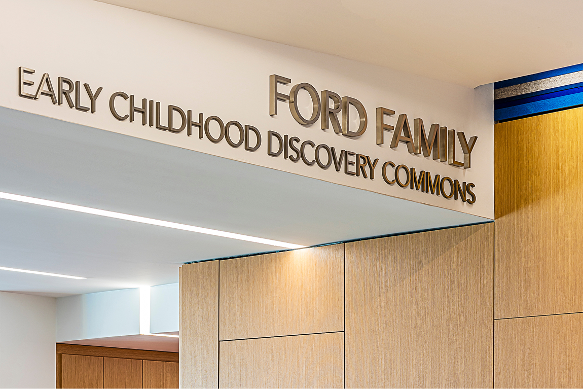 Bronze dimensional letters reading 'Ford Family Early Childhood Discovery Commons' mounted above a wood-paneled entryway with recessed ceiling lighting.
