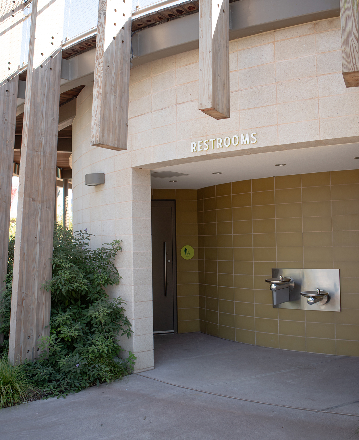 Kaleidoscope Park public restroom exterior entrance with white dimensional letter sign, lime green ADA door placard, stainless steel dual drinking fountains, tan tile wall, and wood post pavilion structure, Frisco, Texas