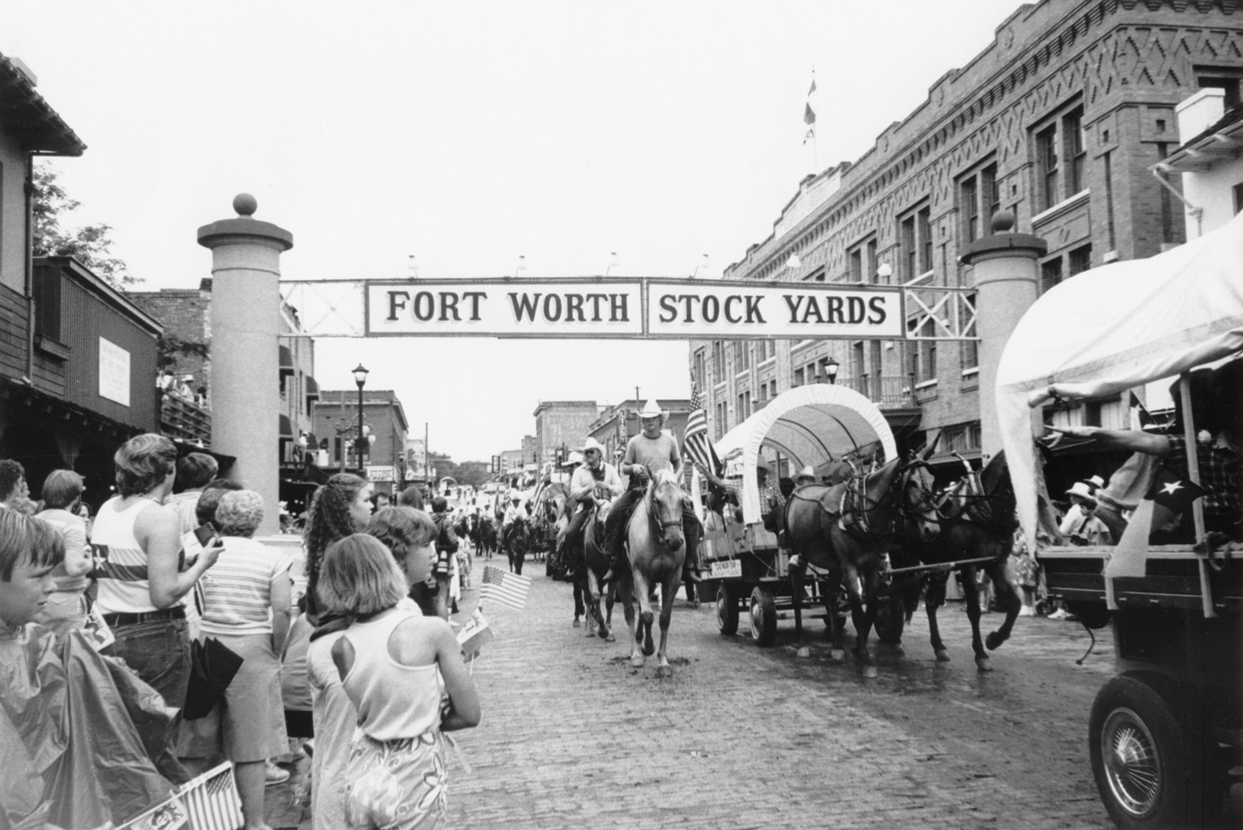 A procession of horse-drawn covered wagons and cowboy-hatted men on horseback travels down a brick road, passing beneath a sign that reads “Fort Worth Stock Yards” in all capital letters. A crowd watches from the sidewalk, with several people holding American flags. On either side of them, historical buildings line the street. The photo is in black and white.