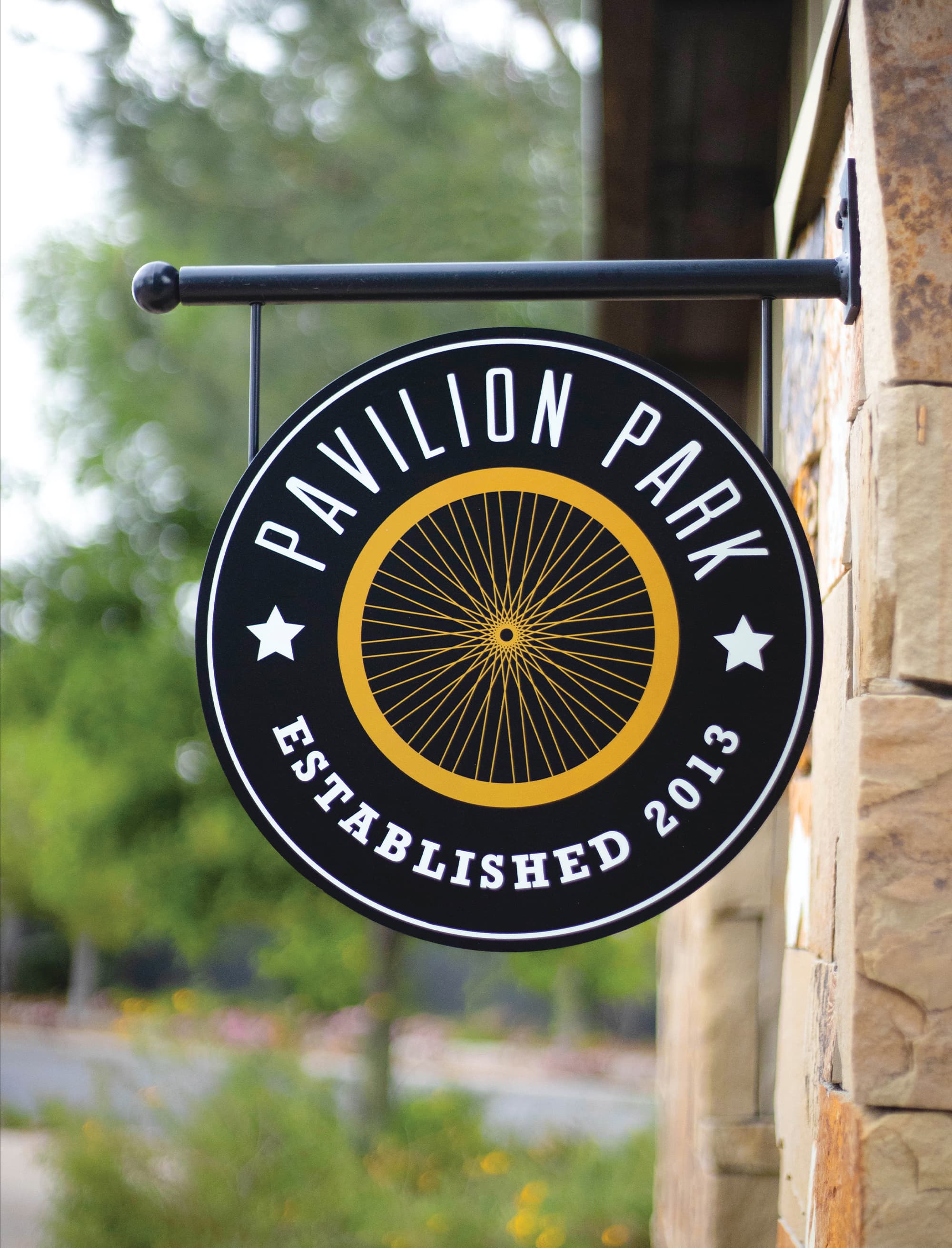 Round black hanging sign reading 'Pavilion Park, Established 2013' with a yellow bicycle wheel graphic and white stars, mounted on a black bracket against a stone wall.