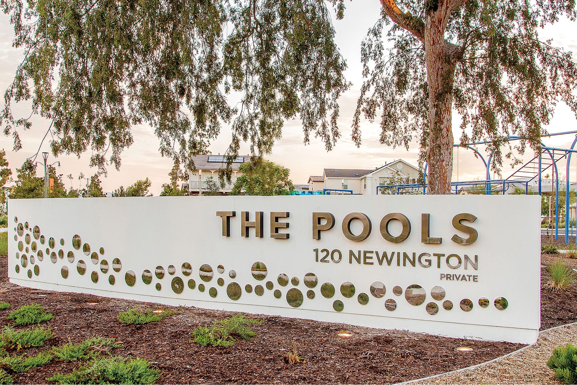 Architectural entrance monument sign for The Pools at 120 Newington Private featuring bronze dimensional letters and decorative circular cutout bubble pattern on a white curved wall in Irvine, California, designed by RSM Design