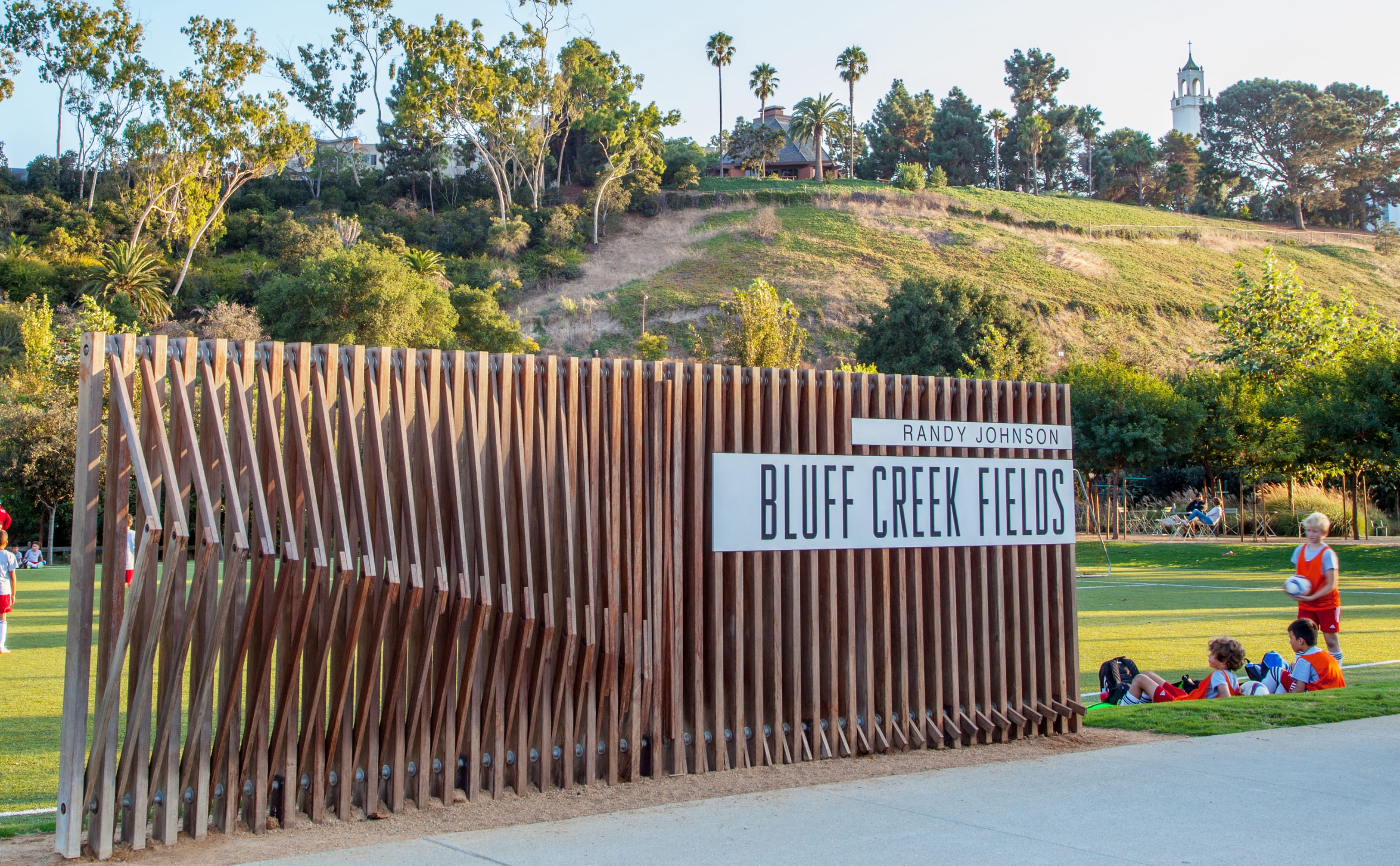 Wood slat monument sign reading "Randy Johnson Bluff Creek Fields," designed by RSM Design, at the edge of a sports field in Playa Vista with children playing soccer nearby.