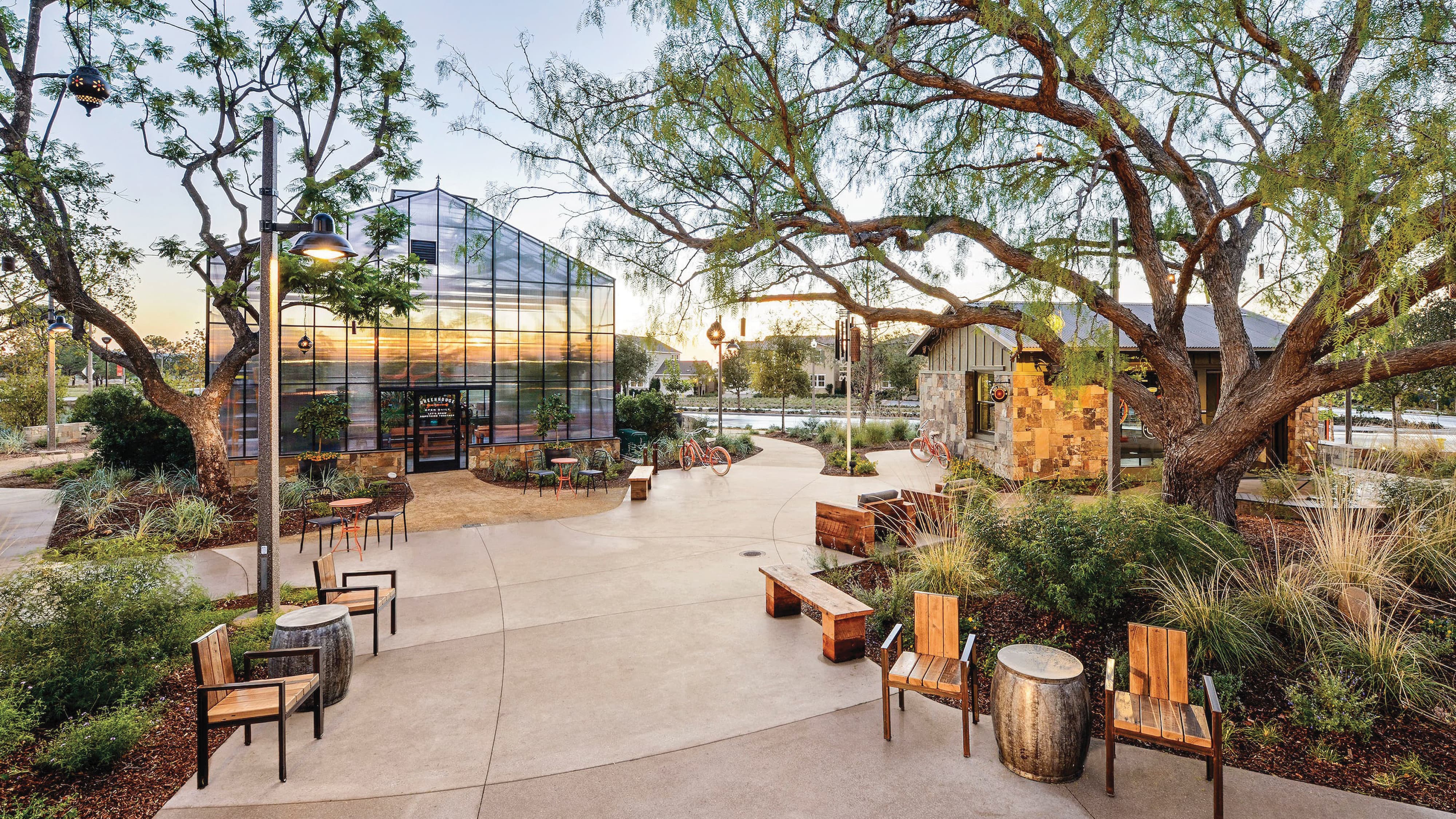 Community gathering plaza at sunset with a greenhouse-style glass building, a stone kiosk, wooden chairs and benches around barrel side tables, orange bicycles parked on a winding path, and mature trees shading the space