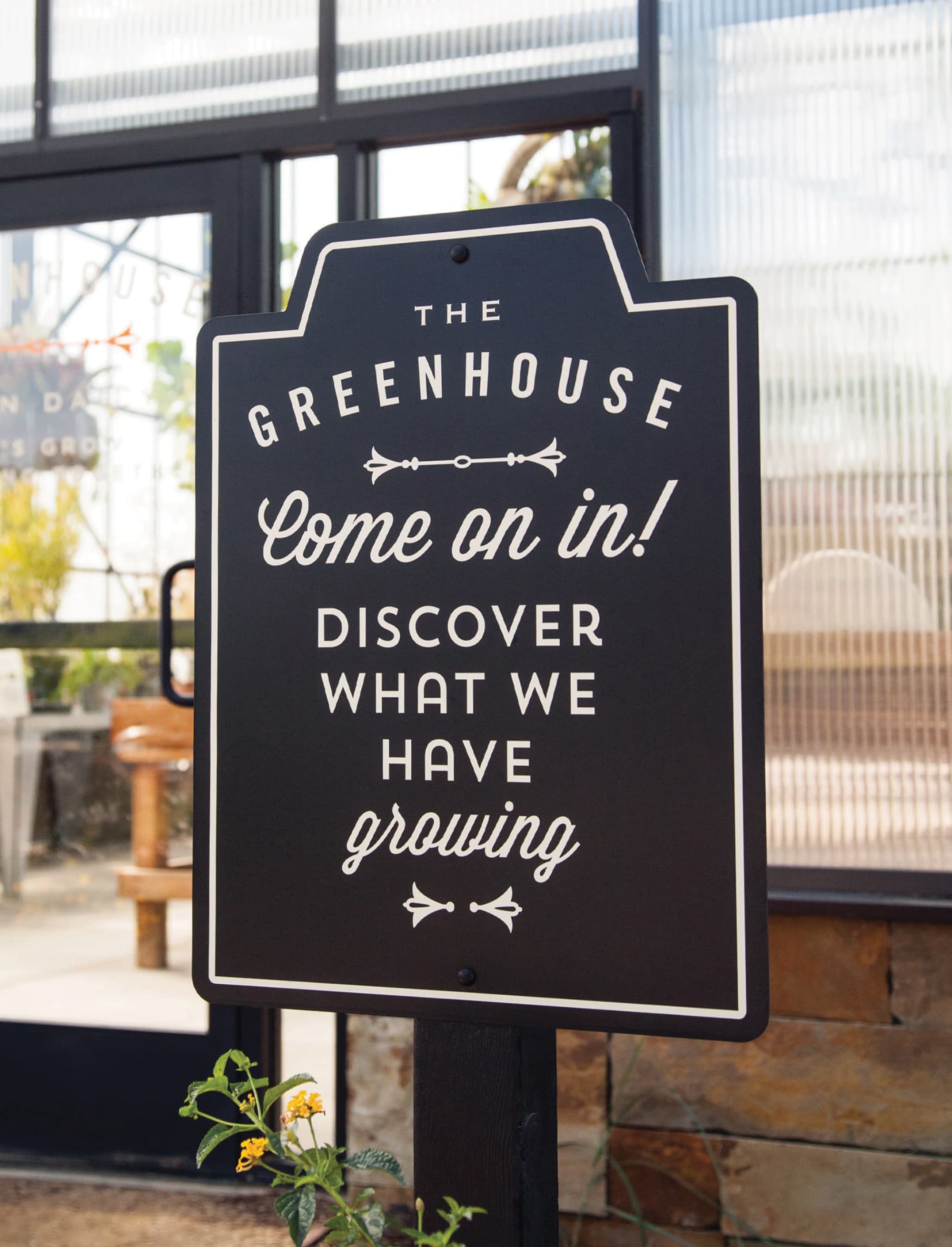 Vintage-style black and white sign reading 'The Greenhouse, Come on in! Discover what we have growing,' mounted on a post in front of the glass greenhouse entrance, with a small flowering plant beside the post.