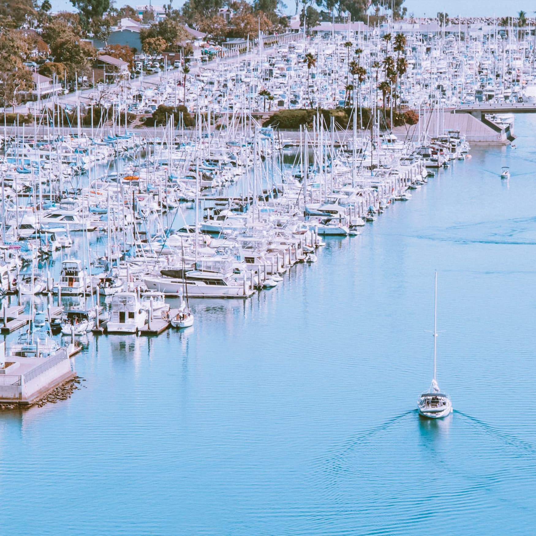 A boat sailing through the main lane at Dana Point Harbor with the current waterfront retail and restaurants in the background.