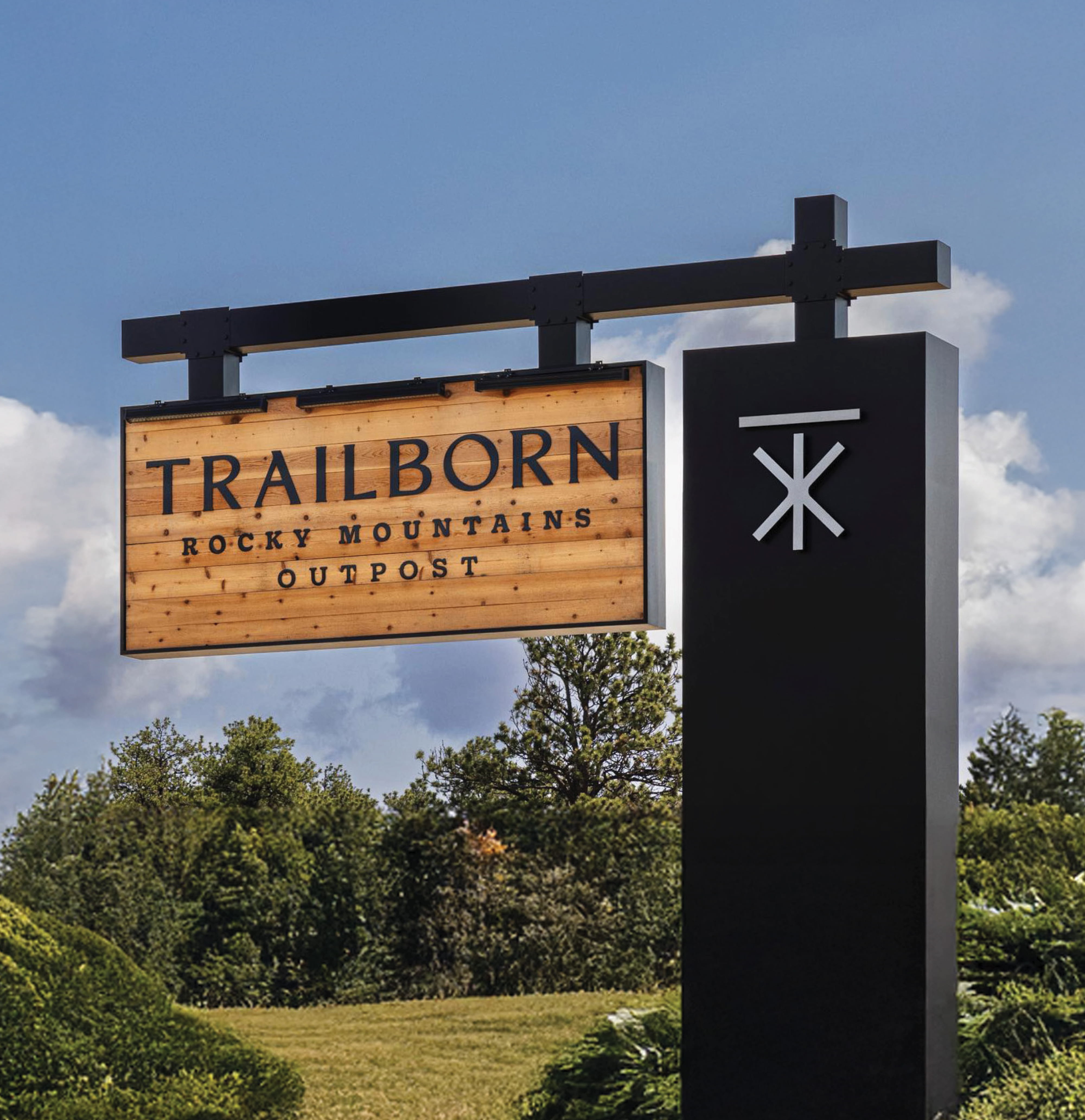 Trailborn Rocky Mountains Outpost monument sign featuring a natural wood panel with routed lettering suspended from a black steel post-and-arm structure with a dimensional brand icon, set against a blue sky and pine tree backdrop, showcasing signage and branded environment design for a hospitality environment by RSM Design.