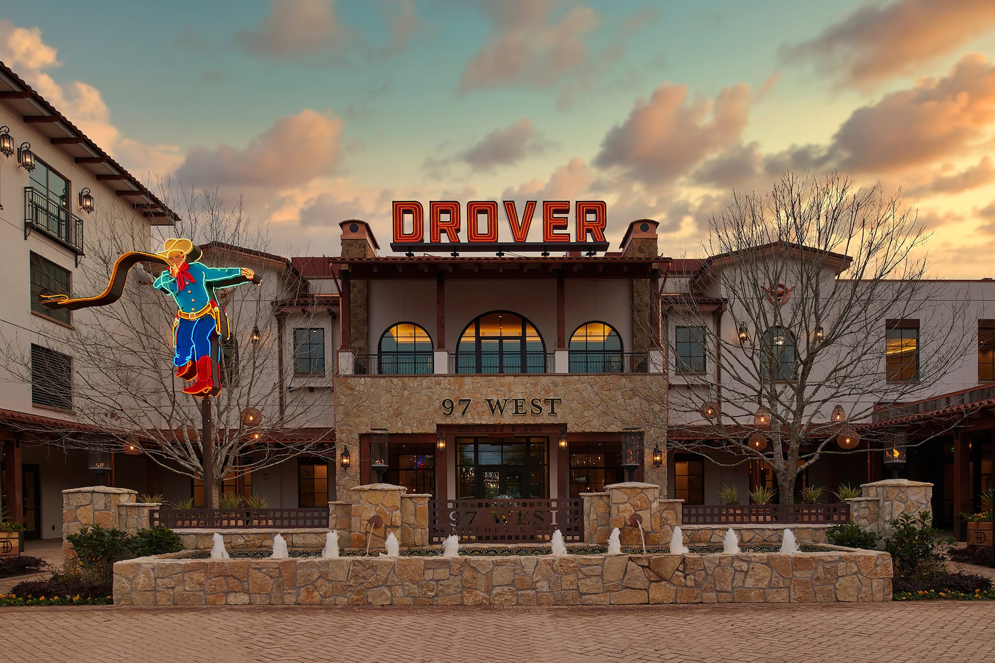 97 West Kitchen and Bar entrance at Hotel Drover in Fort Worth Stockyards, Texas, featuring a glowing neon cowboy sign, red Drover rooftop marquee, limestone facade, arched windows, and courtyard fountains at sunset