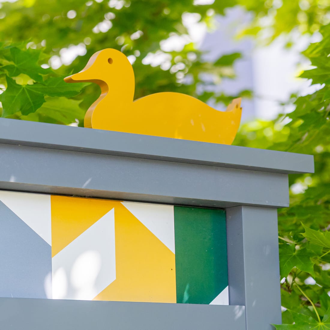 Playful yellow duck cutout topping a gray sign panel with geometric yellow, white, and green graphics at Mountain View Village in Riverton, Utah — environmental graphic design and placemaking by RSM Design.