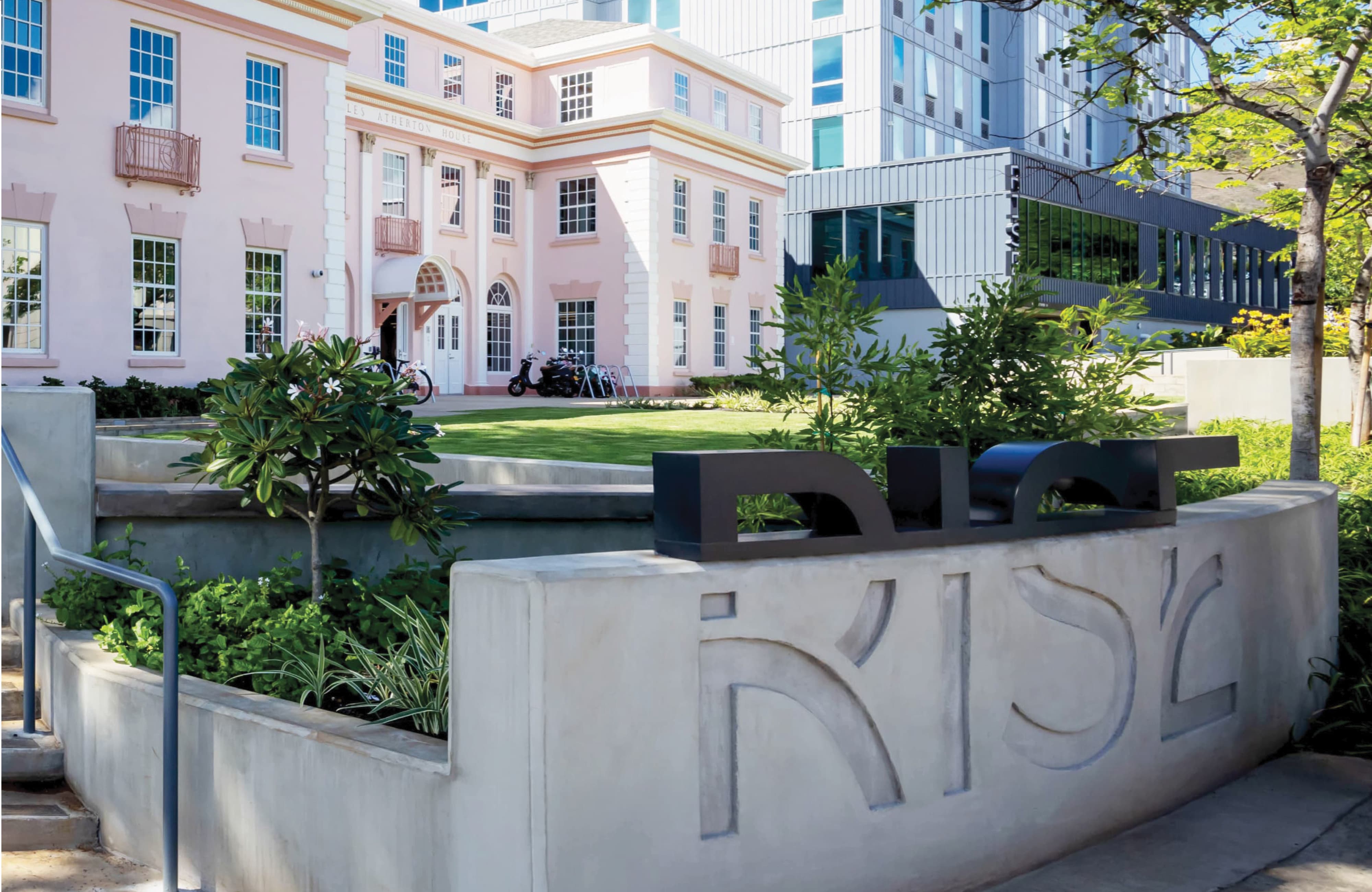 Concrete wall engraved with 'RISE' topped by matching dimensional dark letters, overlooking a courtyard lawn with the historic pink Charles Atherton House and a modern high-rise dormitory in the background.