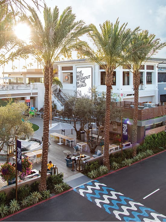 Aerial view of the outdoor plaza at 2nd & PCH in Long Beach, California, featuring a painted chevron crosswalk graphic, branded 2nd & PCH banner signage on light poles, a large-scale tenant wall graphic, and an activated pedestrian gathering space lined with palm trees and outdoor seating — showcasing RSM Design's placemaking, experiential graphics, and environmental graphic design for a coastal waterfront retail and mixed-use destination.