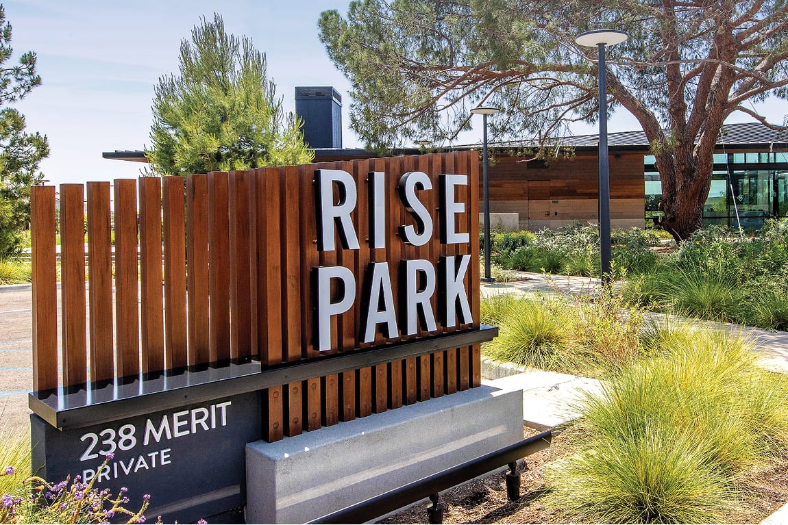 Architectural monument sign for Rise Park featuring white dimensional letters mounted on a warm wood slat panel with a dark metal base, located at 238 Merit Private in Irvine, California, designed by RSM Design
