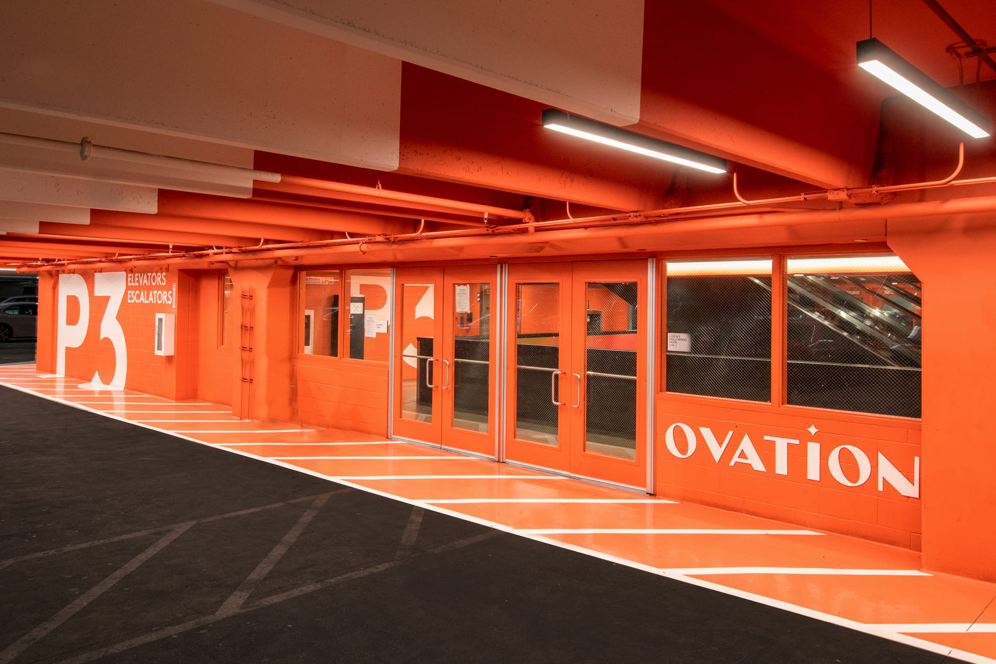 Ovation Hollywood P3 parking garage level with bold all-over orange branded environment, large-scale white "P3" and "OVATION" typography, and elevator and escalator access doors