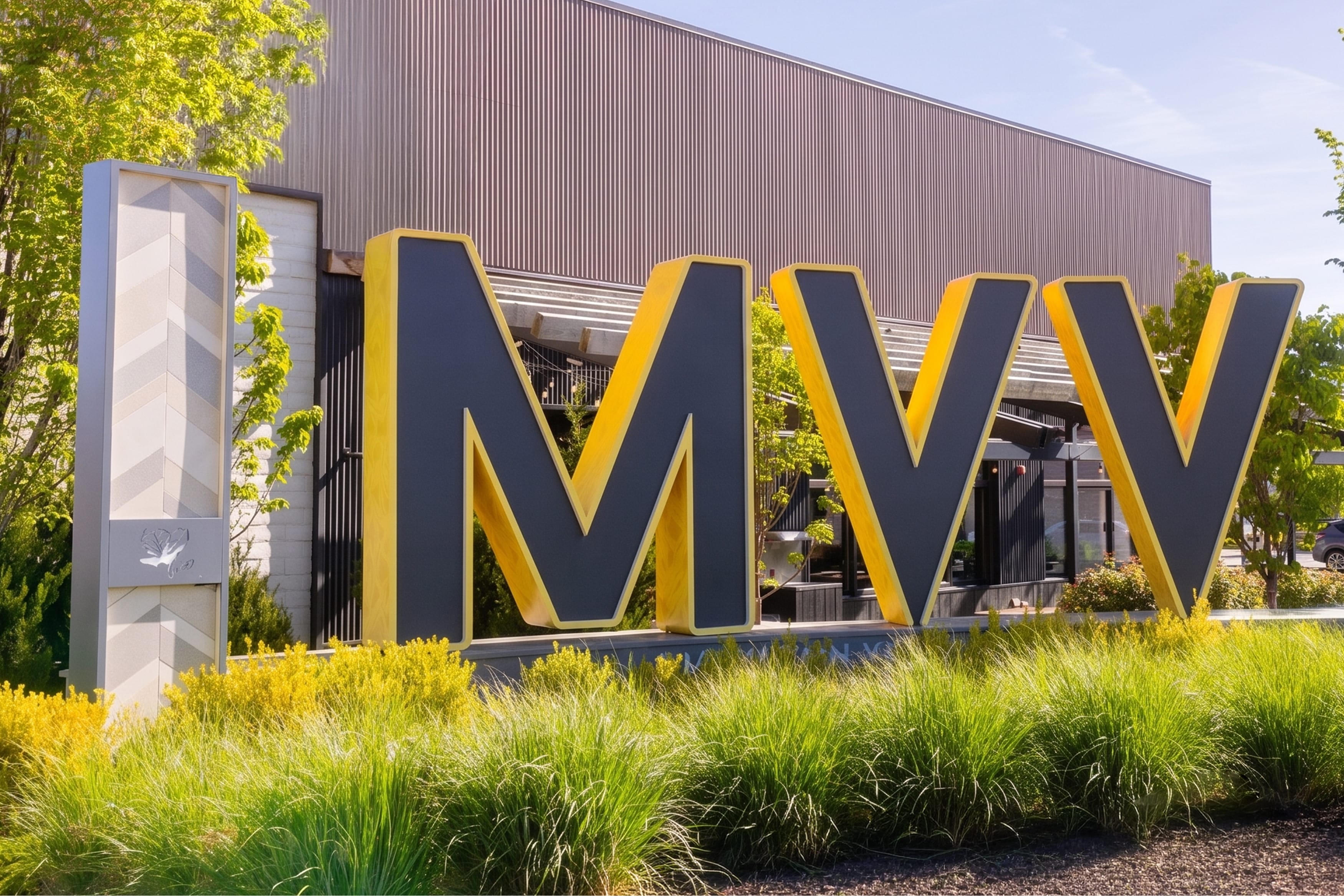 Oversized dimensional "MVV" monument letters in navy blue with yellow-lit sides, set among ornamental grasses at the Mountain View Village entrance in Riverton, Utah — environmental graphic design and monument signage by RSM Design.