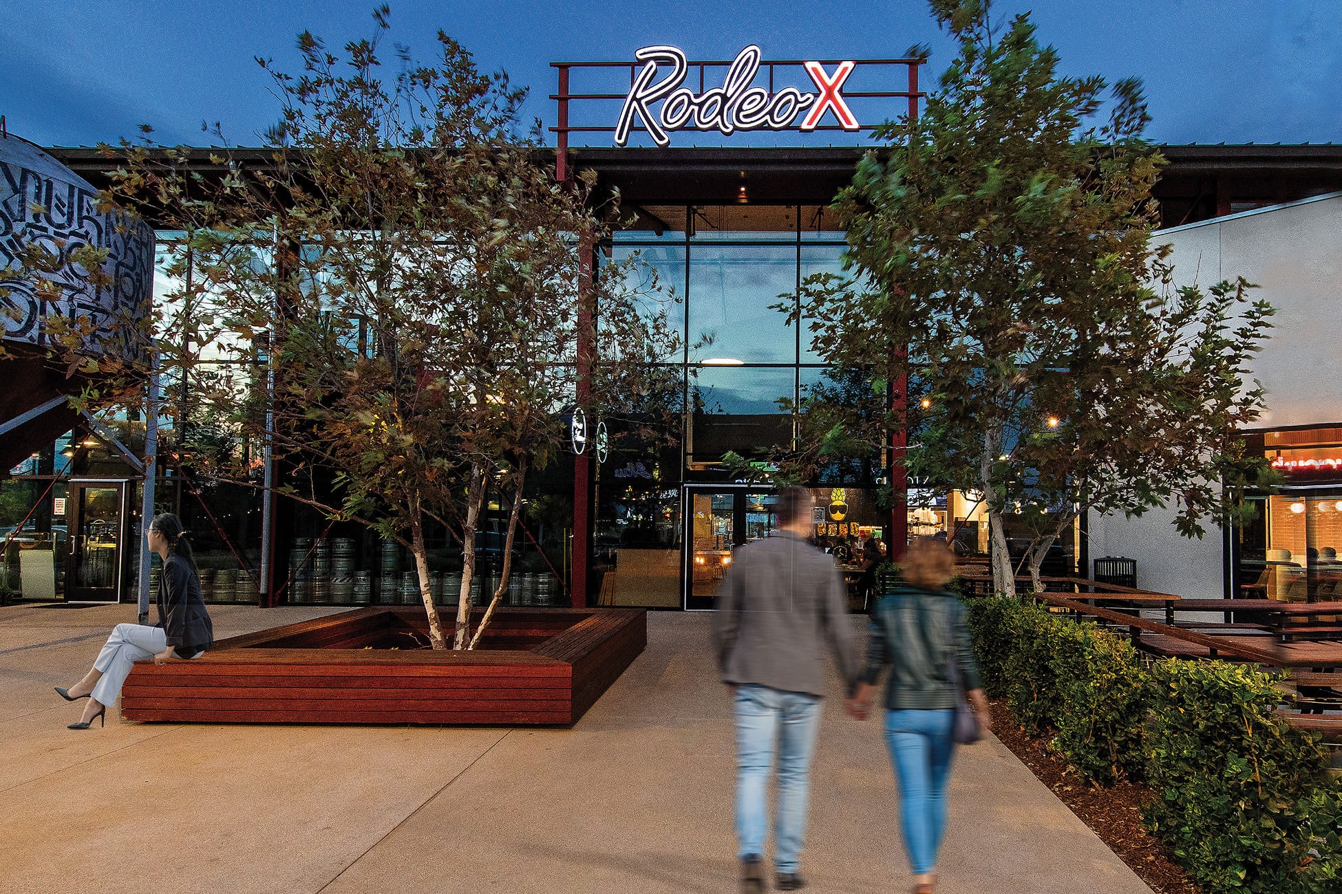 Twilight view of the illuminated 'Rodeo X' sign atop a glass-walled building, with a couple holding hands walking toward the entrance and a woman sitting on a wood planter bench nearby.