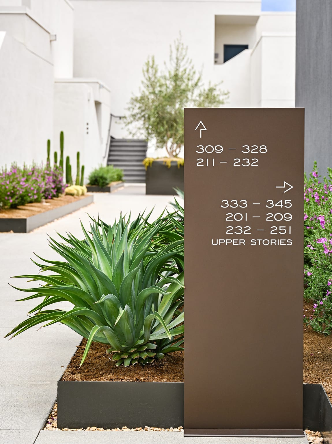 Bronze freestanding wayfinding totem at Thompson Palm Springs directing guests to room ranges and upper stories, set among agave, flowering desert shrubs, and columnar cacti with the white stucco courtyard and stairs in the background.