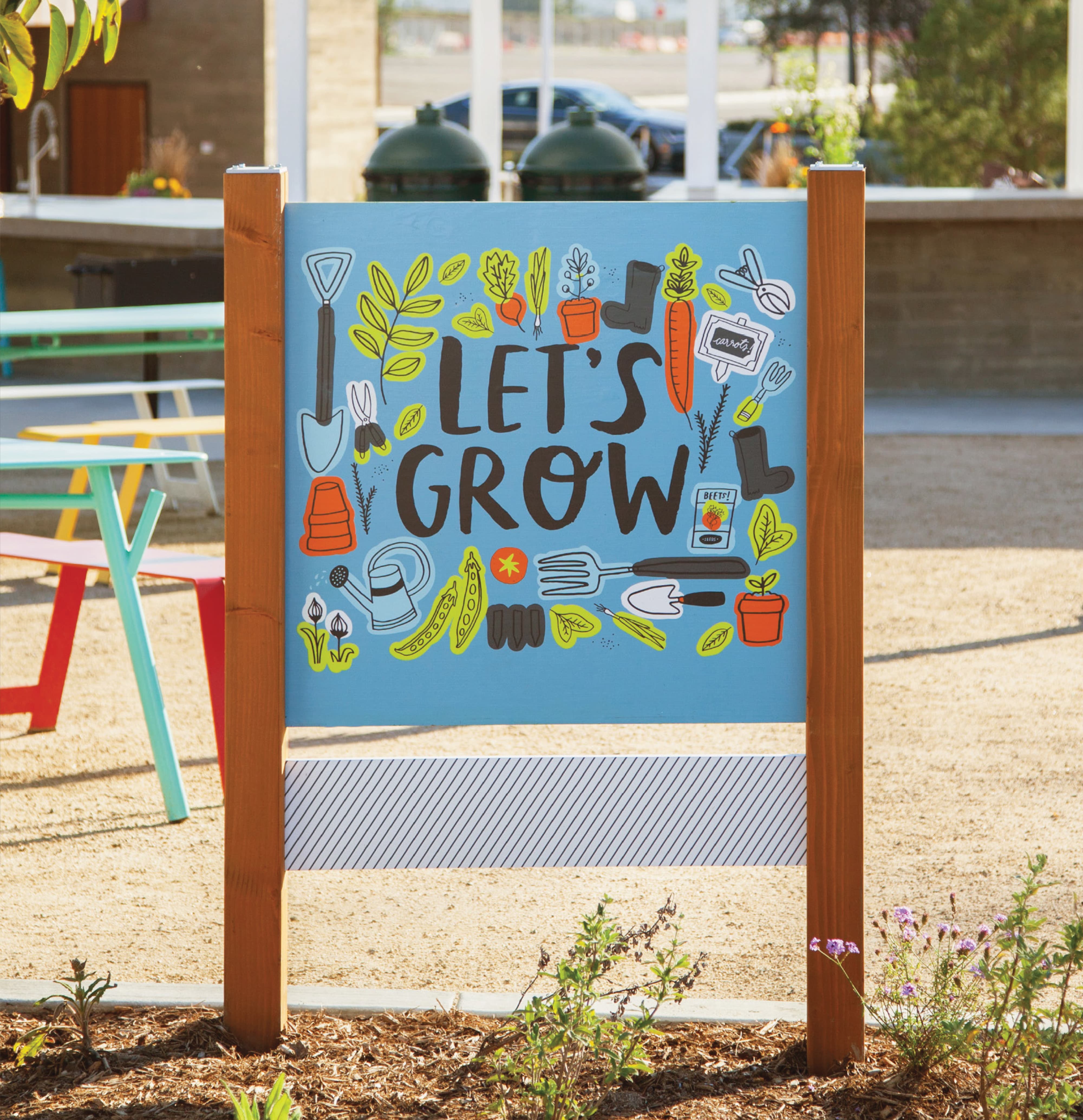 RSM Design experiential graphics and placemaking signage for FivePoint at Great Park Neighborhoods in Irvine, California, featuring a close-up of a colorful illustrated wood post sign reading "Let's Grow" surrounded by hand-drawn garden tool and vegetable iconography on a sky blue background, set within a community garden space with colorful outdoor furniture visible alongside, showcasing how environmental graphic design, specialty graphics, art programs, and placemaking use illustrated storytelling, playful typography, and community-centered messaging to create joyful, human-centered branded environments for residential community and park markets.