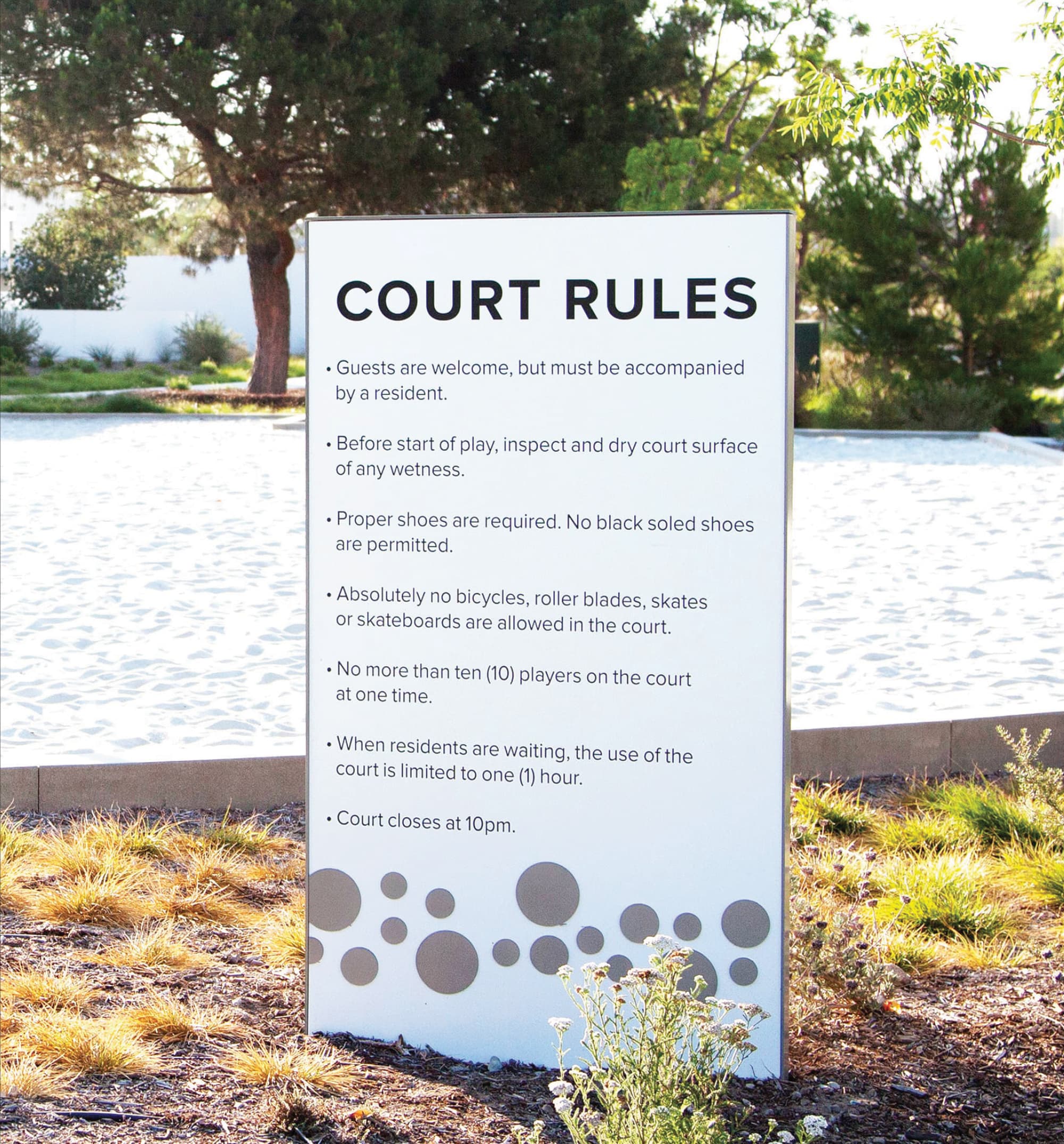 Court Rules sign listing guidelines for guests, footwear, and player limits, positioned in landscaped grasses in front of a light-colored sport court, with trees in the background.