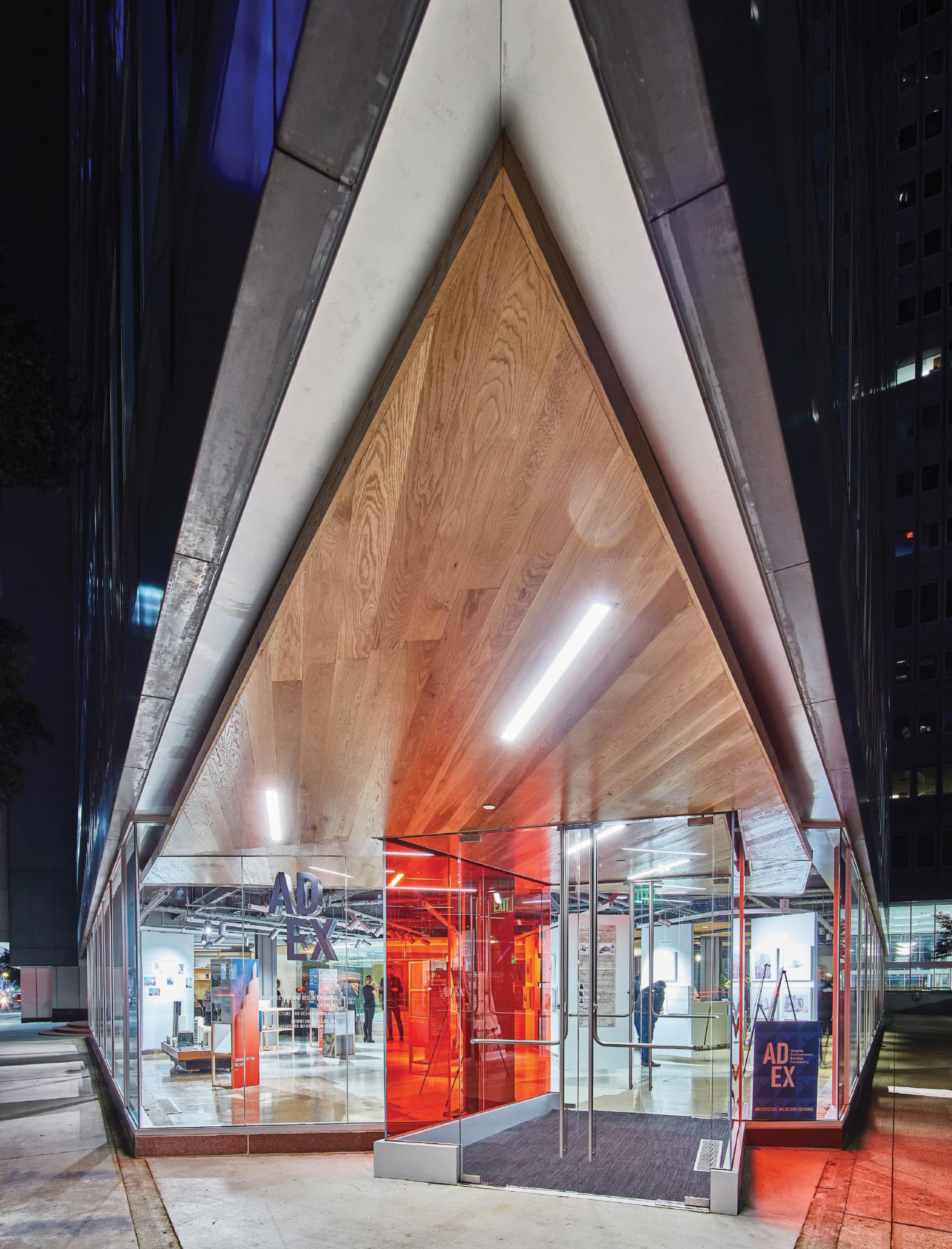 RSM Design entry signage and environmental graphic design for the Architecture and Design Exchange in downtown Dallas, featuring a dramatic nighttime view of the ADEX identity signage and branded red door panels glowing within a soaring triangular wood-clad canopy entry portal on a landmark modernist glass curtain wall building, with visitors visible inside the illuminated interior gallery space, demonstrating how wayfinding and signage design, visual identity, experiential graphics, and placemaking create a powerful, human-centered sense of arrival for cultural, civic, and urban streetscape markets.