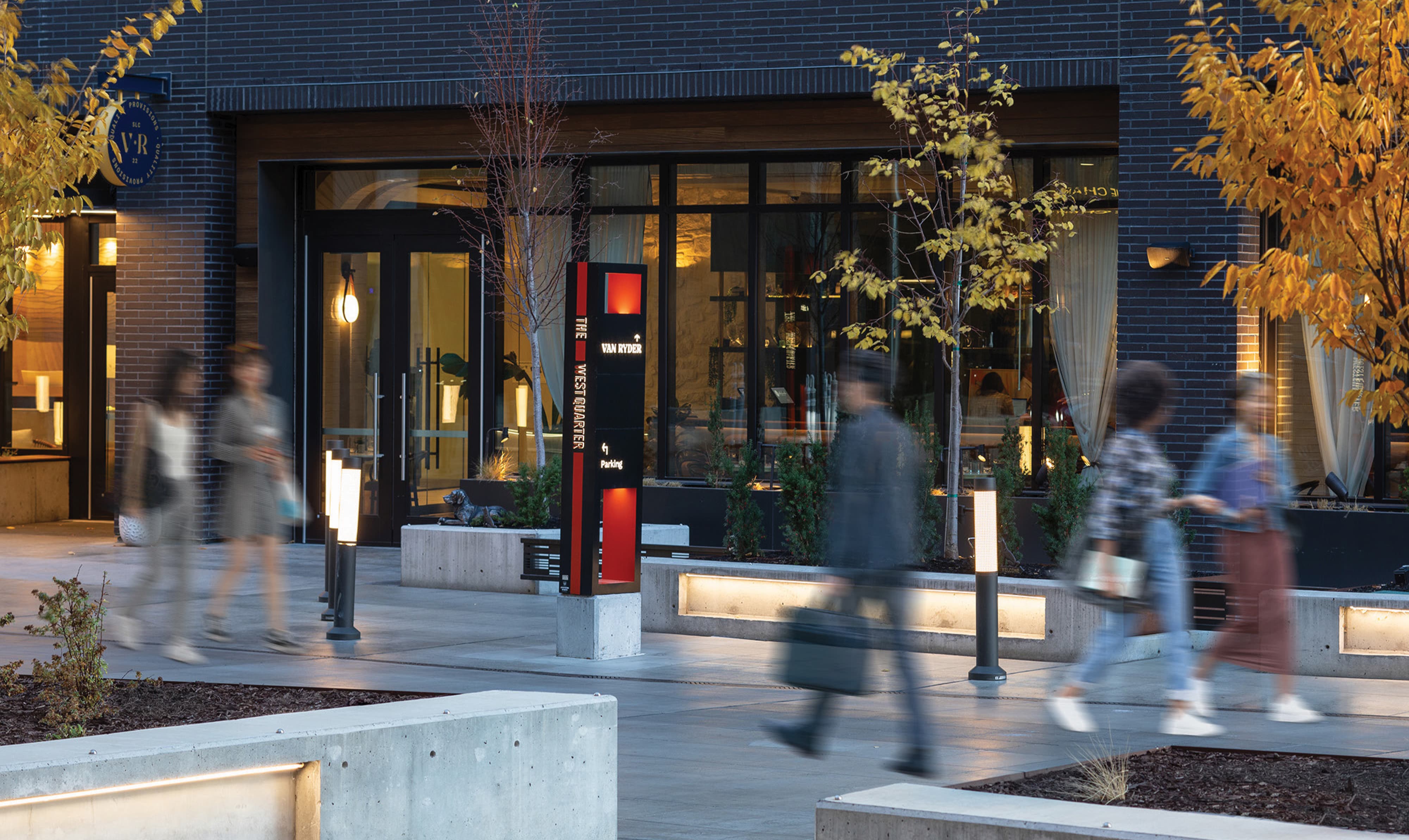 RSM Design pedestrian wayfinding pylon and branded environment for The West Quarter in Salt Lake City, featuring a tall black and red illuminated directional sign identifying Van Ryder and parking navigation set within an activated evening plaza surrounded by motion-blurred pedestrians, bollard lighting, concrete seating, and autumn foliage, with The Charles and additional tenant storefronts visible in the background, demonstrating how wayfinding and signage design, environmental graphic design, master sign program, and placemaking create intuitive, human-centered navigation experiences for mixed-use hospitality, food and beverage, residential community, and urban streetscape markets.