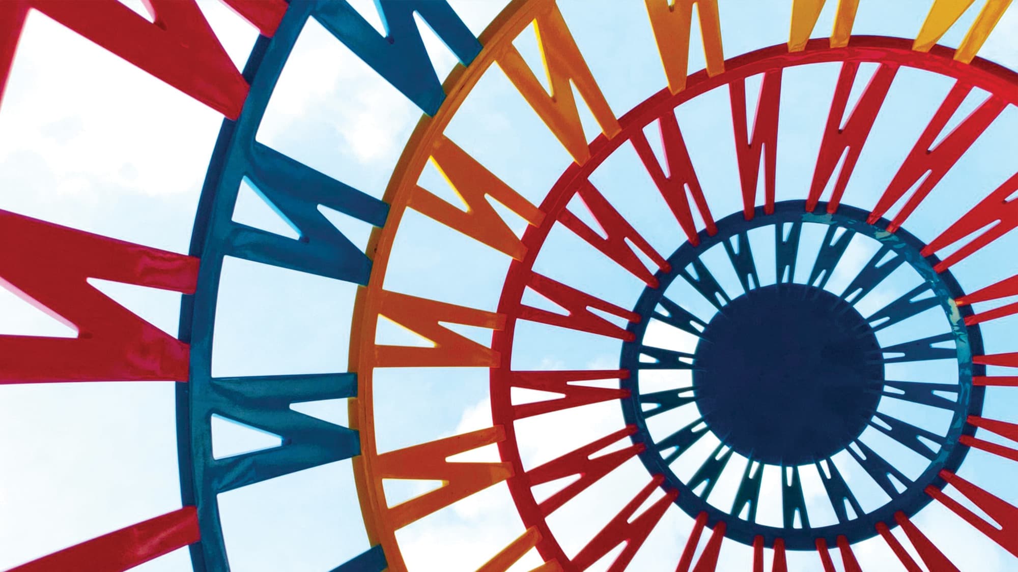 View looking up at concentric red, blue, orange and yellow rings whose zigzag spokes converge on a navy center, against a pale blue sky.