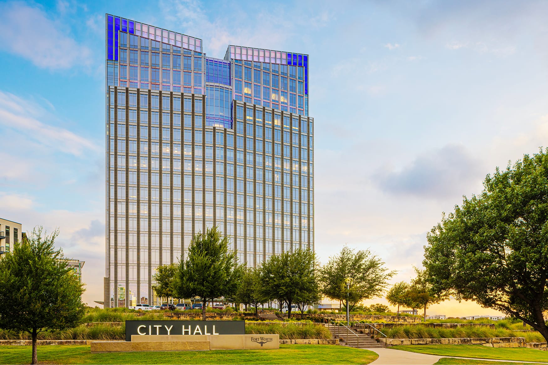 Fort Worth City Hall glass tower exterior at golden hour featuring illuminated blue and purple upper facade, City Hall monument sign with Fort Worth longhorn logo, landscaped terraces, and tree-lined entry plaza