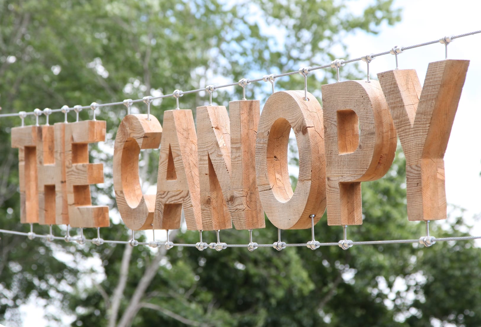 Suspended reclaimed wood dimensional letters spelling The Canopy hanging from steel cable at Summit Bechtel Reserve, creative outdoor placemaking signage design by RSM Design
