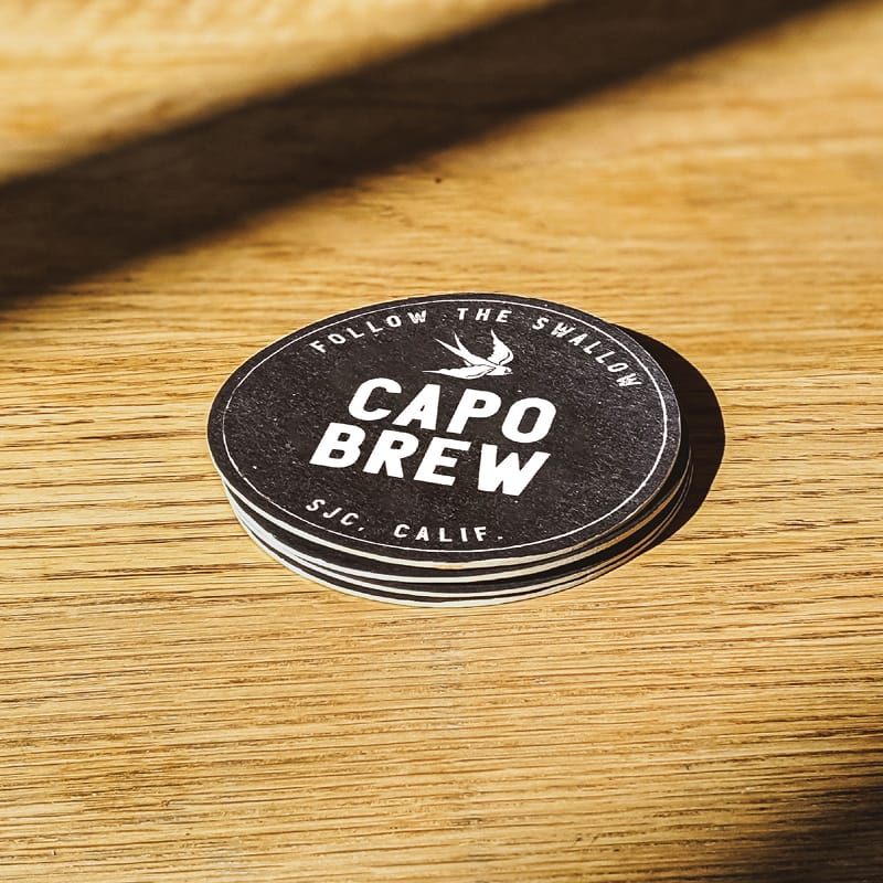 Stack of round black Capo Brew coasters with swallow icon and "SJC, Calif." text on a wood table, brand identity design by RSM Design