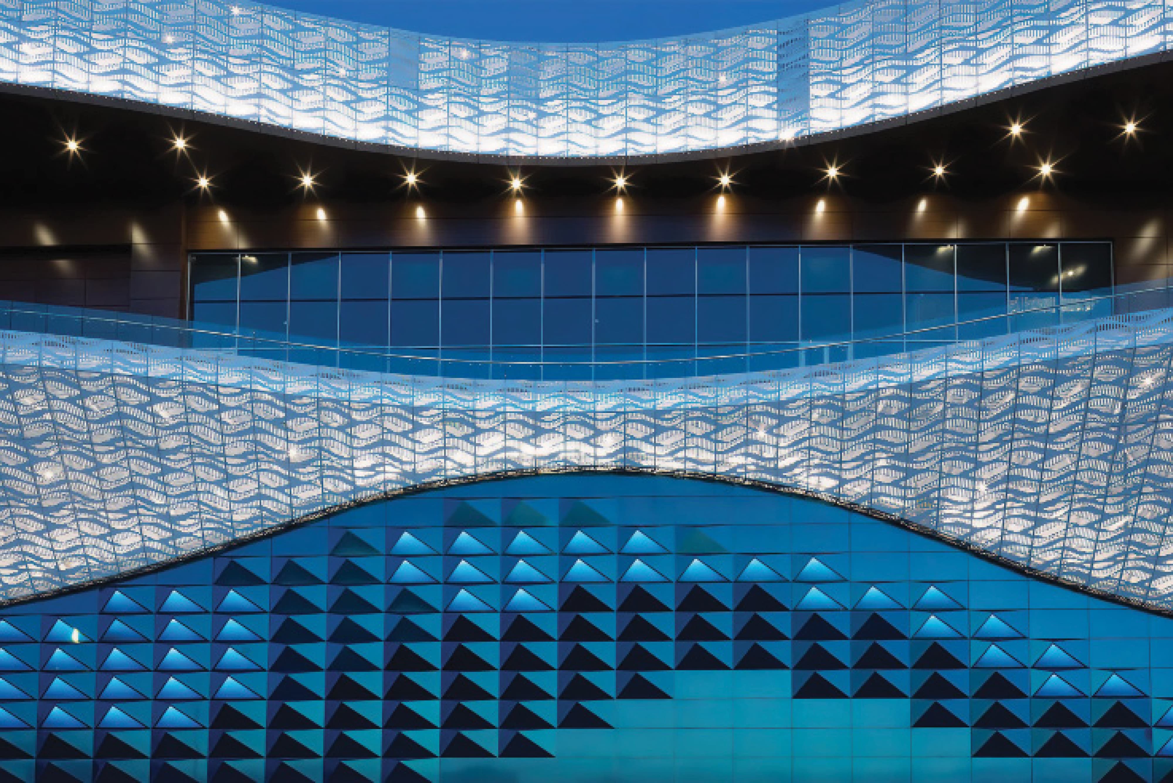 Large-scale interior architectural facade featuring illuminated wave-patterned perforated metal panels and a geometric blue triangulated glass wall at an international retail and mixed-use destination — RSM Design experiential graphics, specialty graphics, branded environments, art programs, and placemaking for retail, mixed-use, and international destinations.